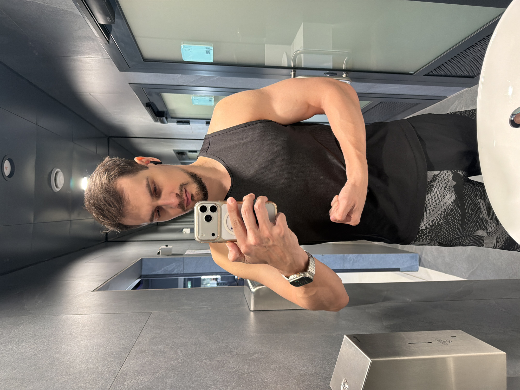 A man is taking a selfie in a public restroom. He is wearing a black tank top and black shorts. He is flexing his right bicep. He is holding an iPhone in his left hand, and wearing a silver Apple Watch on his left wrist. The restroom has gray tile walls and floors. There is a mirror behind the man, and a sink and a metal soap dispenser in the foreground. A window is visible behind the man. The location is in the city of Krasnoyarsk, Russia.