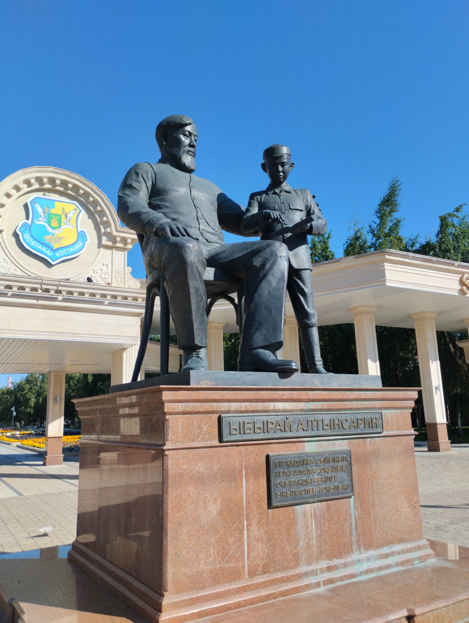 A bronze statue of two figures, an adult male and a young boy, sits on a granite pedestal. The adult, seated, wears a suit and has a beard. The boy, standing, wears a uniform and holds a book. Above them, in the background, is a cream-colored building with classical columns and an emblem featuring two birds and a shield with the year "1879". The emblem also includes text in Cyrillic, reading "QOSTANAI KOSTANA". This monument is located in Kostanay, Kazakhstan, likely in a public square or park given the surrounding paved area, landscaping with yellow and orange flowers, and trees. The sky is clear and blue.