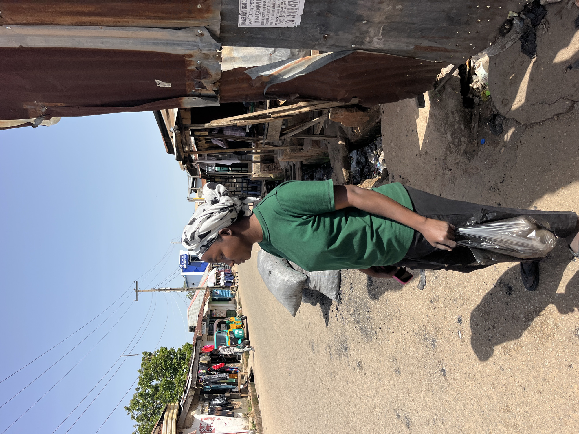 A medium shot captures a woman standing on a dusty street in a bustling market area, her profile facing left. She wears a green t-shirt, black pants, and a patterned headscarf. In her right hand, she clutches a clear plastic bag, and a pink mobile phone is visible in her left hand. Behind her, several large, dark sacks are piled on the ground. The street is lined with small shops, some with corrugated metal roofs and walls, others with open fronts displaying merchandise like clothing. A bright yellow three-wheeled auto-rickshaw is parked on the street further down. The ground is a mix of dirt and scattered debris. The sky above is clear and blue. The scene appears to be in an outdoor urban market environment.