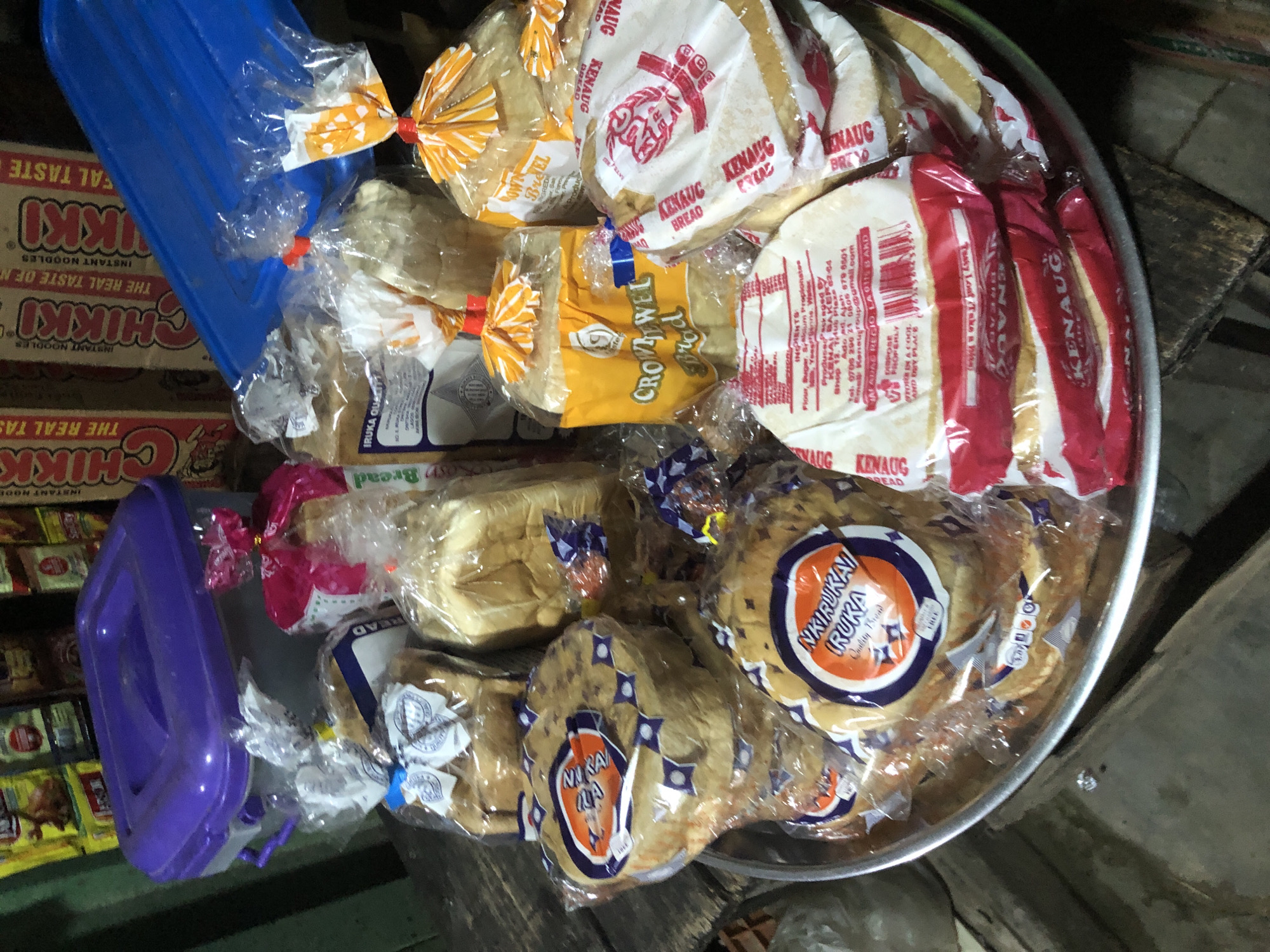 The image depicts a display of packaged bread loaves and rounds on a circular metal tray. The bread is primarily presented in clear plastic packaging, many of which are adorned with colorful labels. Several loaves are round and flat, with labels indicating "Nkiruka Ruka Quality Bread." Other loaves are more elongated, with labels such as "Crown Jewel Bread." Some packages have red and white striped labels that read "KEnaug Bread." Behind the tray, several more loaves are visible, some in yellow packaging. In the background, a purple plastic container with a lid sits next to a blue plastic object. Boxes of "Chikki Instant Noodles" are also visible in the background, suggesting a market or store setting. The wooden surface beneath the tray and the overall lighting suggest an indoor environment, possibly a market stall in Ado, Nigeria, given the presence of local bread brands.