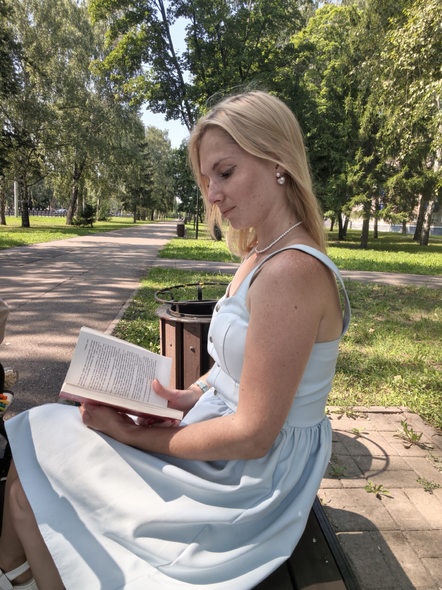 A blonde woman sits on a park bench, reading a book outdoors. She is wearing a light blue dress and pearl earrings. The park appears to be well-maintained with paved walkways and trees lining the path. The scene suggests a peaceful, leisurely day in a city park.