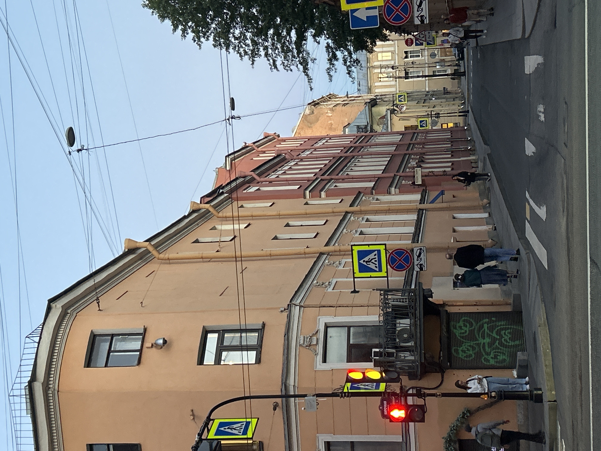 The image depicts a street scene in a European city, possibly Saint Petersburg, Russia, judging by the architecture and street signs. The perspective is from street level, looking down a street lined with multi-story buildings. The buildings exhibit classical architectural styles with ornate facades and numerous windows. The street is paved and has pedestrian crossings marked with white lines.
In the foreground, a traffic light is red, indicating a stop. Pedestrian crossing signs, featuring a blue square with a white silhouette of a person walking, are visible on poles. A streetlamp hangs over the street, supported by wires.
Several individuals are present on the sidewalk and street. One person, wearing a backpack, stands near the traffic light. Further down the street, a group of people walks along the sidewalk. One person is on their phone, while others are walking in pairs.
The buildings are painted in various colors, including peach, red, and a lighter yellow further down the street. Some buildings have balconies. Graffiti is visible on a metal shutter on one of the buildings. Trees are visible in the background, suggesting a green urban environment. The sky is clear and appears to be twilight or early morning. The overall impression is of a quiet urban street during a transitional time of day.