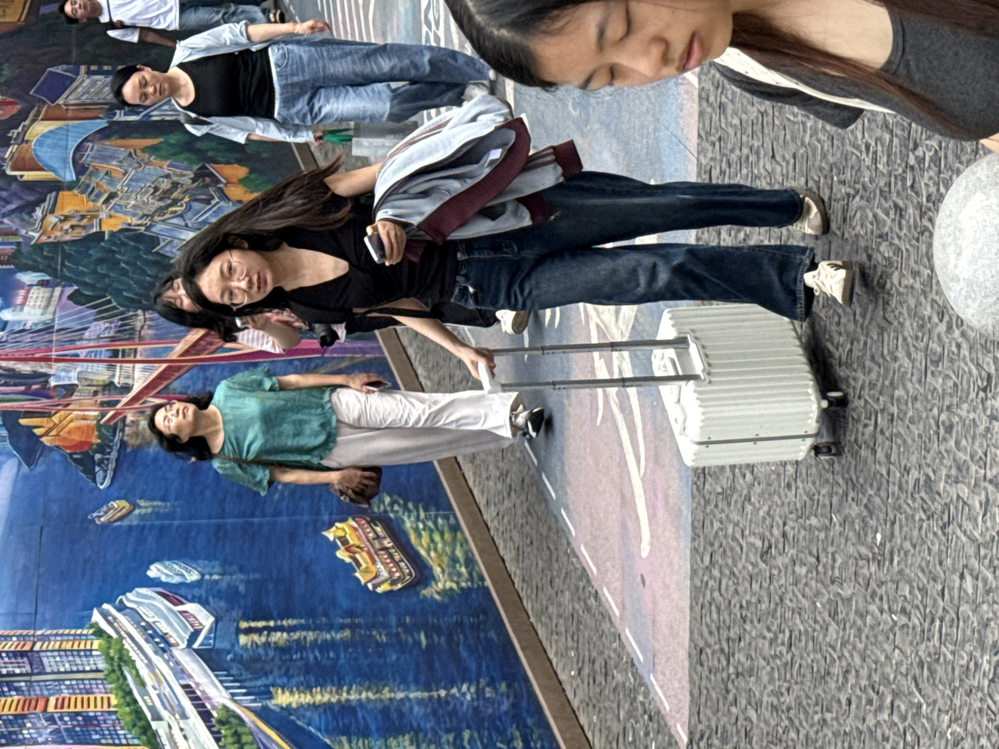 The image depicts a group of people posing in front of a large mural. The location appears to be a street in Yuzhong District, China, based on the style of buildings and murals.
The mural shows an aerial view of a cityscape, with buildings, roads, and water. Several people are in the foreground. An Asian woman with glasses, wearing a black shirt and jeans, is holding a suitcase. Another woman in a teal shirt and white pants is positioned to her left, also interacting with the mural. Another Asian woman in jeans and a black shirt is in the background, posing by laying horizontally against the mural. Other people in the background are also interacting with the mural. The ground appears to be cobblestone or a similar paved surface. The lighting suggests it is daytime.