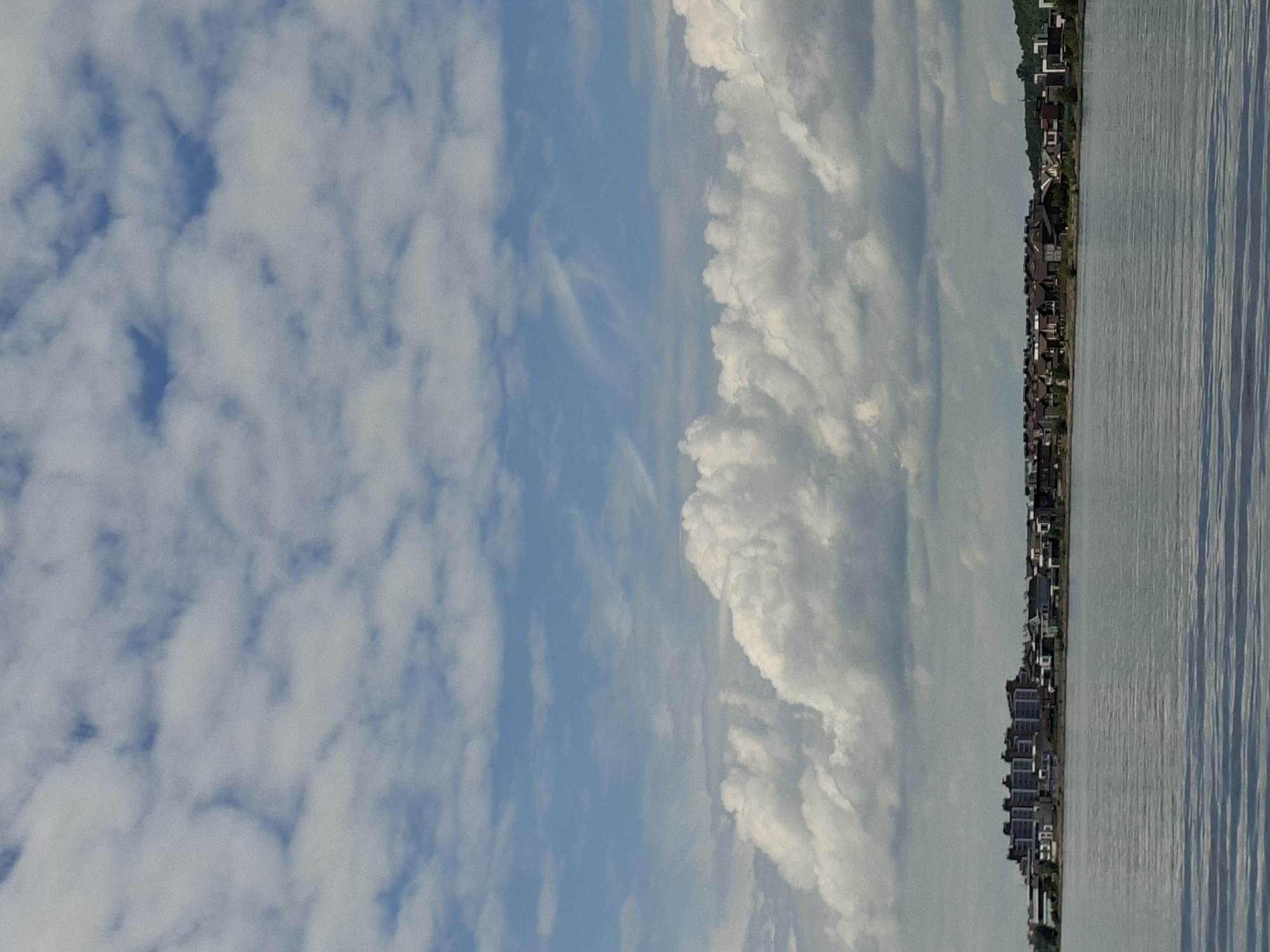 The image depicts a wide, calm body of water with a distant shoreline lined with buildings. The sky is filled with a mix of fluffy white clouds and patches of blue. The shoreline features a dense cluster of multi-story apartment buildings on the left, followed by a stretch of smaller houses with dark roofs. The water reflects the sky and the buildings, creating a somewhat hazy and subdued scene. The overall impression is of a suburban or semi-urban waterfront. This scene is consistent with the appearance of the city of Kazan, Russia, along the Volga River, where modern apartment complexes and residential areas are situated along the waterfront.