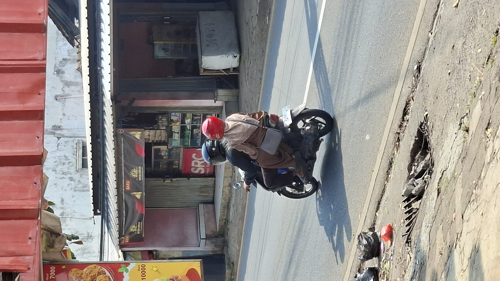 Two individuals are riding a black scooter on an asphalt road. The driver, positioned in the front, is wearing a black helmet. The passenger, seated behind the driver, is wearing a red helmet and a brown hijab, and is holding a gray bag. The scooter is moving away from the camera, heading towards the right side of the image. In the background, there are storefronts with awnings and signs. One sign displays "BARU" and logos. Another sign on the left side of the image features images of fried chicken and text indicating prices like "7000" and "10000". To the right of the storefronts, there is a large, light-colored object covered with a tarp. In the foreground, along the side of the road, there is debris, including fallen leaves, a broken grate over a hole in the ground, and two black trash bags. The scene appears to be in a commercial area.