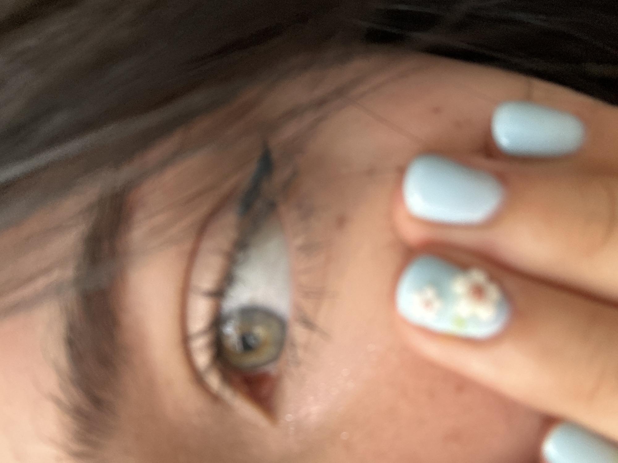 This is a close-up, upward-angled, and slightly blurred medium shot of a person's eye and hand. The eye is looking to the right and has a greenish-brown iris. Dark hair is visible in the upper left corner of the frame, partially obscuring the eye. The person's hand is in the foreground, with pale blue nail polish on their fingernails. One fingernail features a delicate floral design with small white flowers and touches of pink and green. The skin tone appears fair. The background is out of focus, showing hints of light and shadow. There is no visible text. The scene suggests a personal moment of self-care or preparation, with the focus on the detail of the nails and the eye. The lighting is soft and natural, suggesting an indoor setting during daylight.