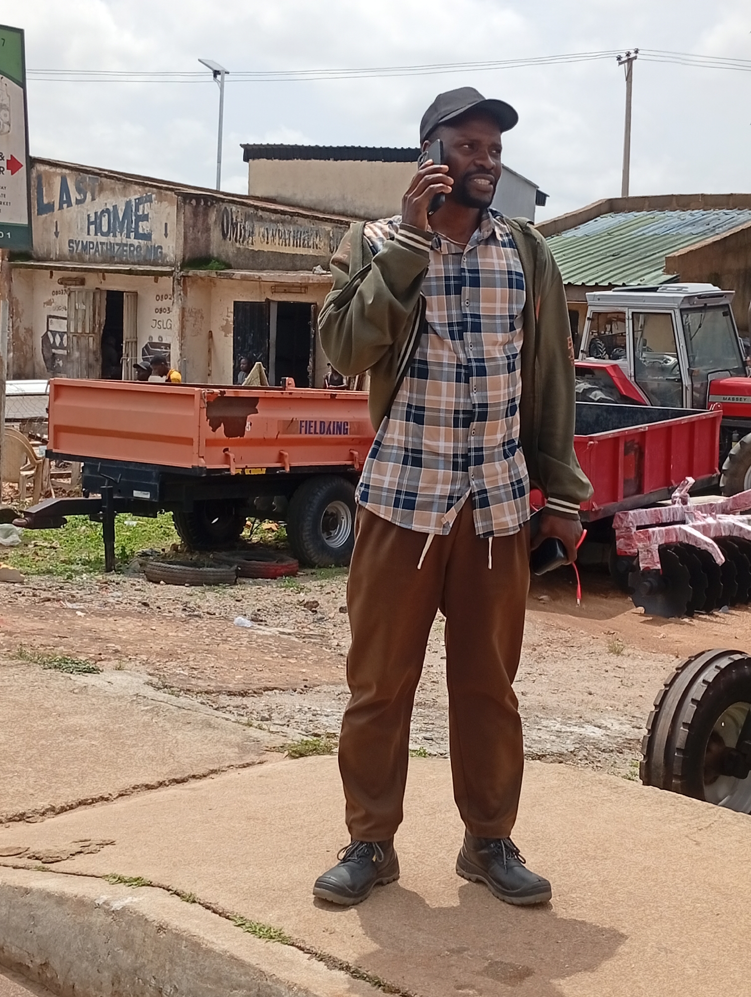 A medium-shot, eye-level outdoor photograph shows a Black man standing and talking on a cellphone. He is wearing a black baseball cap, a plaid button-up shirt, a green jacket, brown pants, and black work boots. The man is holding a black cellphone in his right hand and a black object with a red cord in his left hand. Behind the man is an orange trailer and a red tractor. The background includes buildings, power lines, and a cloudy sky. The scene appears to be in a dusty, rural area.