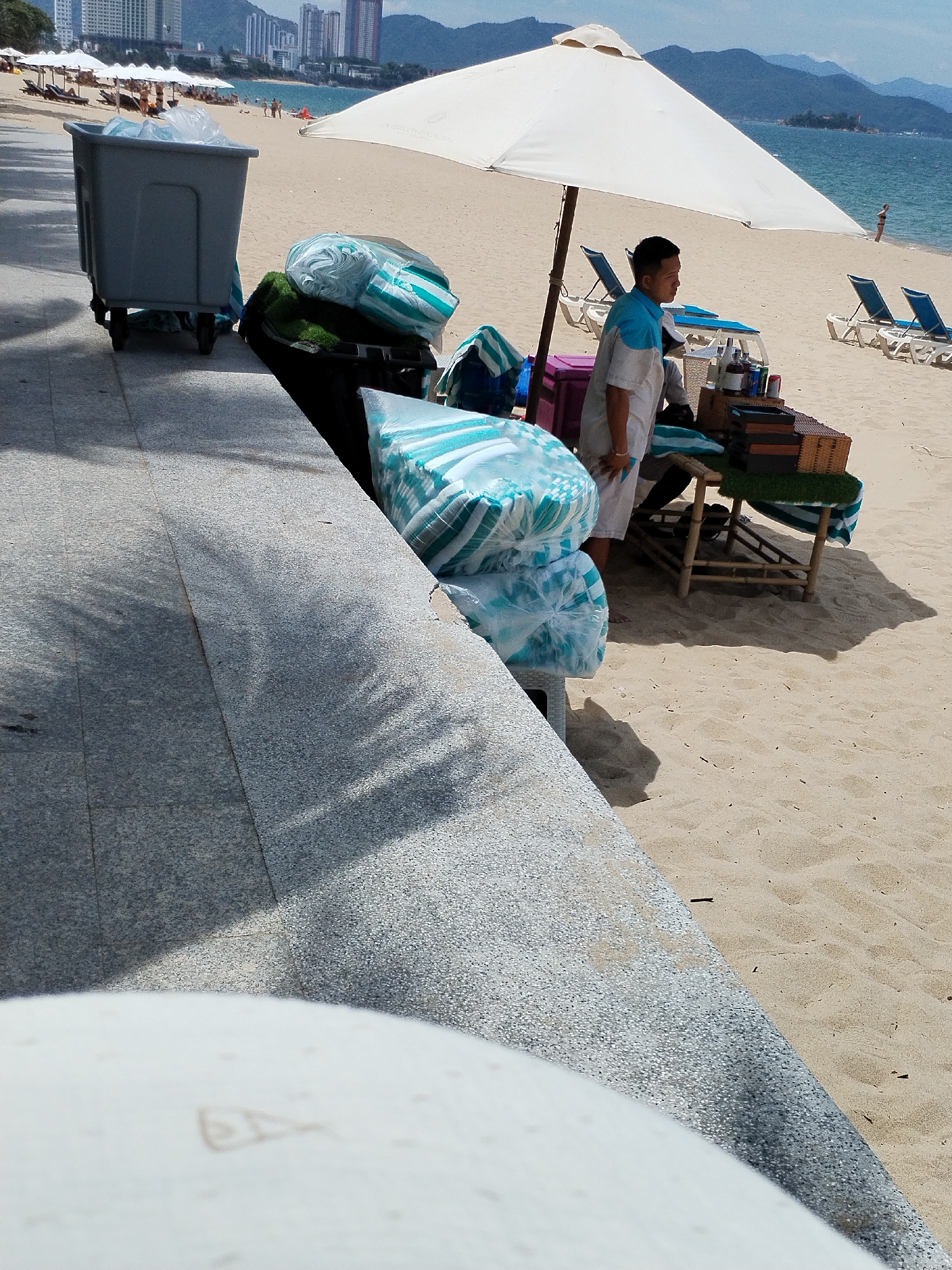 The image shows a beach scene in Nha Trang Ward, Vietnam. A person, likely a vendor, stands under a large white umbrella, which is positioned on the sandy beach. The vendor is wearing a light blue and white uniform. Next to the vendor is a small table made of bamboo, displaying a few items. Behind the umbrella, several beach chairs are visible on the sand. To the left of the scene, a large gray bin with wheels is present, with some rolled-up items, possibly towels or mats, piled next to it and inside the bin. The background features the ocean and a distant cityscape with tall buildings. The overall atmosphere is that of a commercial area on a beach.