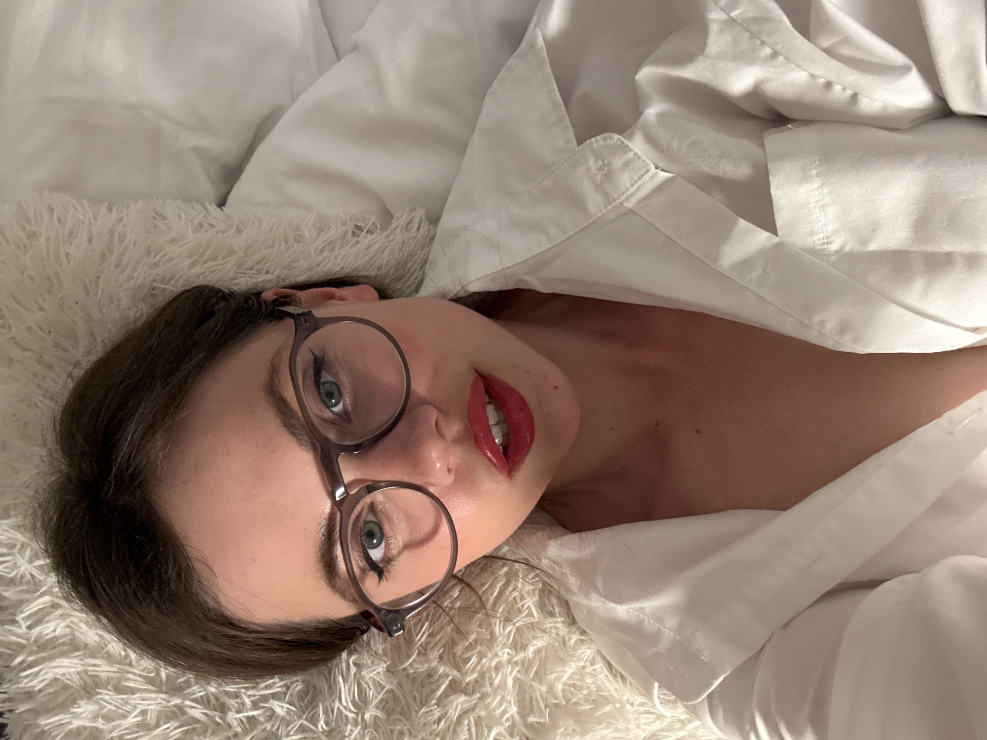 A woman with brown hair, green eyes, and red lipstick, wearing glasses and a white collared shirt, is lying down on a white furry blanket. The background consists of white pillows and the furry blanket. The image appears to be a selfie taken from a high angle. The scene is indoors, and there is no information that places this image in Vrchlabí, Czechia.