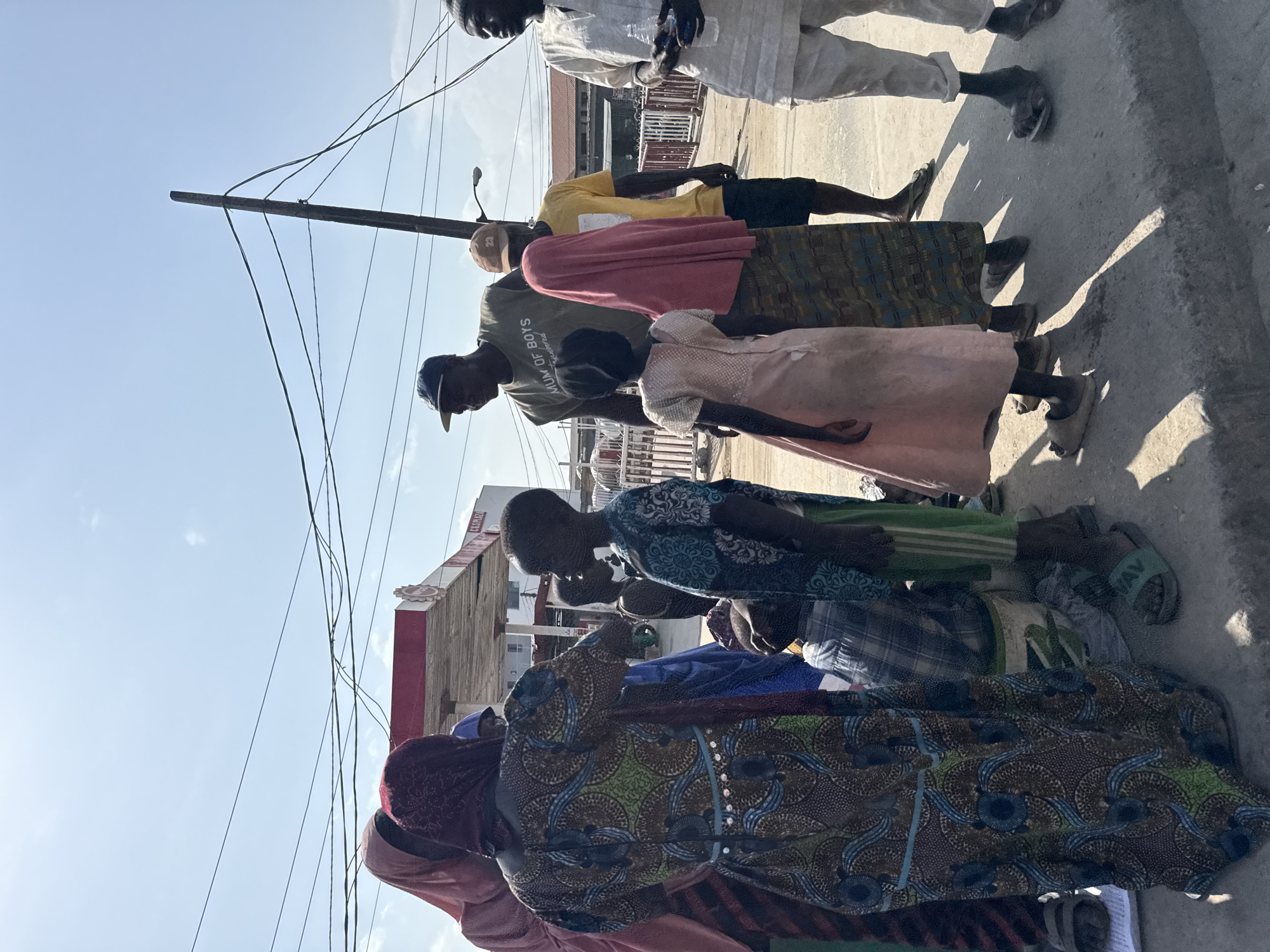 A group of people of varying ages and genders are standing together on a sidewalk in Ado, Nigeria. Most are wearing traditional African clothing made of brightly colored fabric. The group is standing on a concrete sidewalk in front of a building with a red and white sign. An overhead power line crosses above the group. The sky is blue.