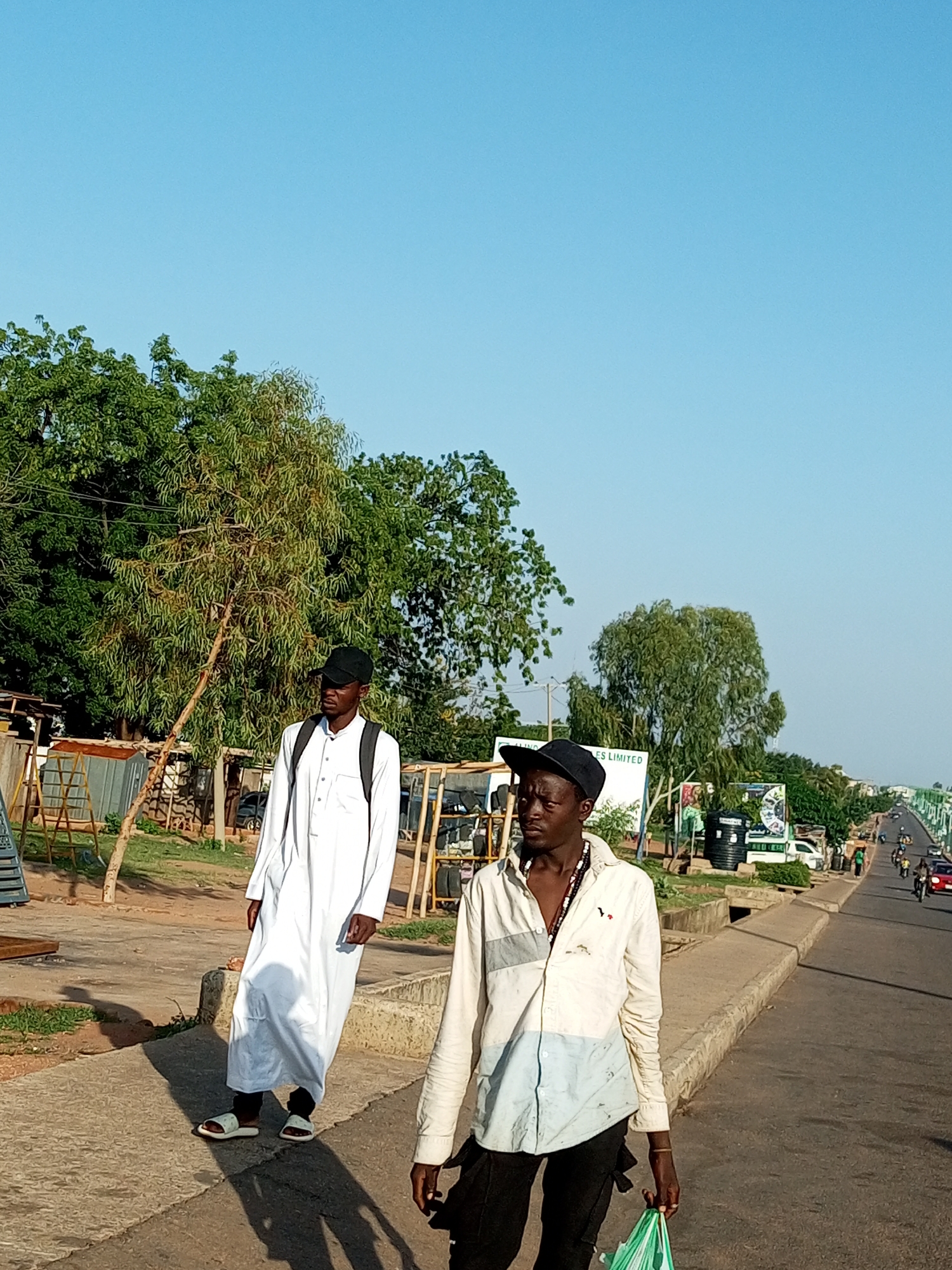 In the city of Garko, Nigeria, two young men are captured in a street scene. The man on the left, facing away from the camera and towards the right, is wearing a white traditional Islamic robe and a black cap. He has a black backpack slung over his shoulders and is walking on a paved walkway. He is wearing white sandals. The man on the right, facing the camera with his head turned slightly to his left, is wearing a light-colored, long-sleeved collared shirt with blue and grey accents and a black baseball cap. He is holding a green plastic bag in his right hand and is wearing dark pants. He has a beaded necklace. They are walking on a paved surface adjacent to a road where traffic, including motorcycles and cars, is visible in the distance. The environment features lush green trees, some with thin trunks, and structures that appear to be market stalls or workshops. The sky is a clear, bright blue. The sun casts long shadows on the ground, indicating it is likely late afternoon. A concrete curb separates the walkway from the road, and a drainage ditch is visible along the side of the road. Several water tanks and signs are present along the roadside.