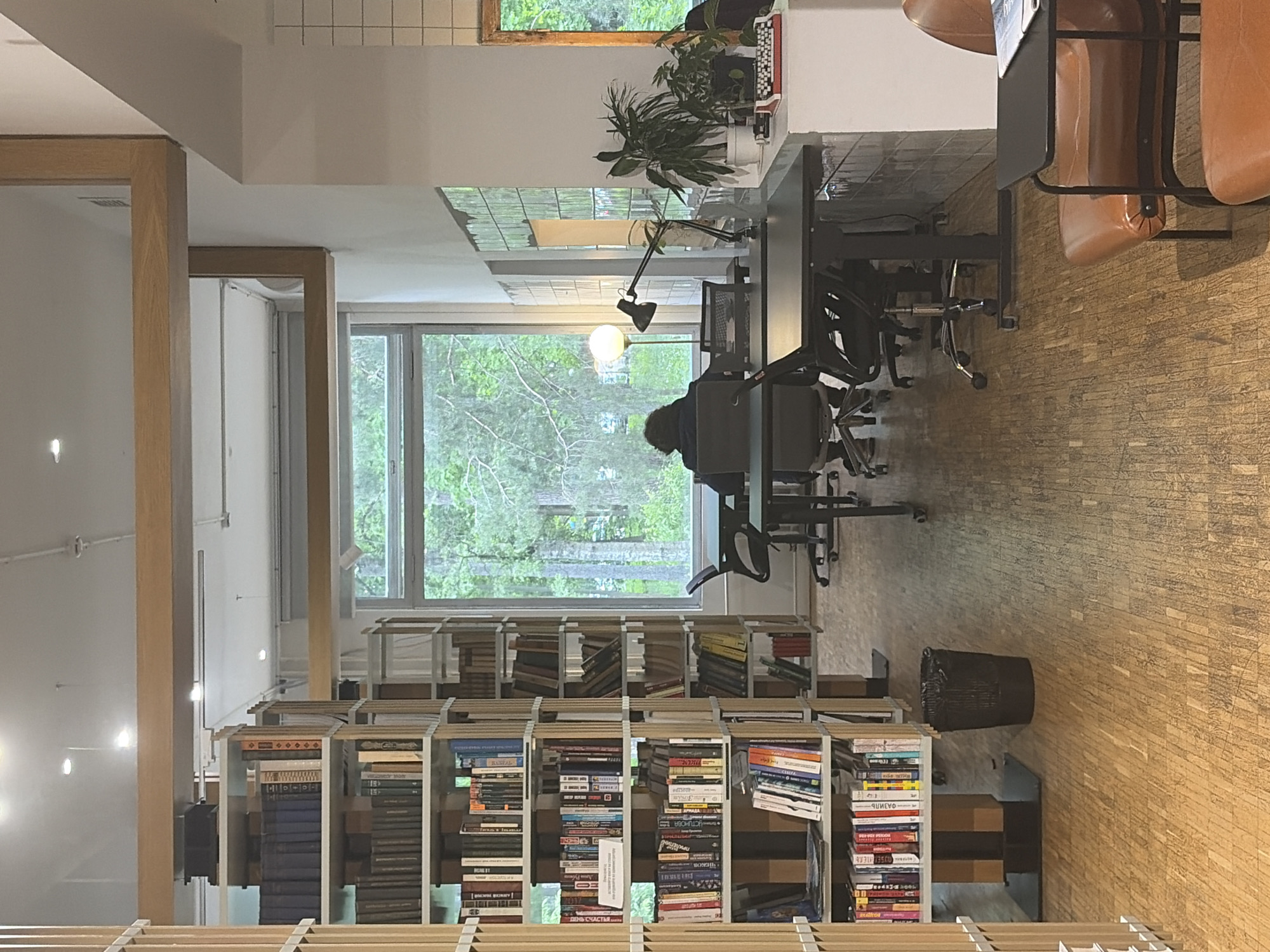 Here is a detailed description of the image:
**Setting:** The photograph appears to be taken indoors, possibly in a library or a shared workspace, with a bookshelf acting as a partition. Large windows look out onto a dense green landscape, suggesting a location near a park or a wooded area.
**People:** The only visible person is seated at a desk with their back to the camera. It is impossible to discern their gender or age from this vantage point.
**Objects:**
* **Bookshelves:** Tall bookshelves filled with books of various sizes and colors act as a room divider.
* **Books:** The shelves are densely packed with books, suggesting a focus on reading or study.
* **Desks and Chairs:** Several desks with office chairs are positioned near the windows, indicating a workspace environment.
* **Lamps:** A modern desk lamp is visible, providing light for the workspace.
* **Plants:** There are plants placed on a higher shelf by the desk, likely for decoration or to improve the atmosphere.
* **Trash can:** A black trash can is near a bookshelf.
**Actions:** The person is seated at the desk, seemingly engaged in work or study. Other actions may be happening out of frame, but the photograph provides no direct information.
**Interactions:** The person is interacting with their workspace, likely using a computer or reading materials. There are no interactions between people visible in the image.
**Environment:** The room appears well-lit due to the large windows, and the interior design emphasizes a modern and comfortable workspace. The overall environment seems conducive to concentration and productivity. The city and the neighborhood of this location is unknown.