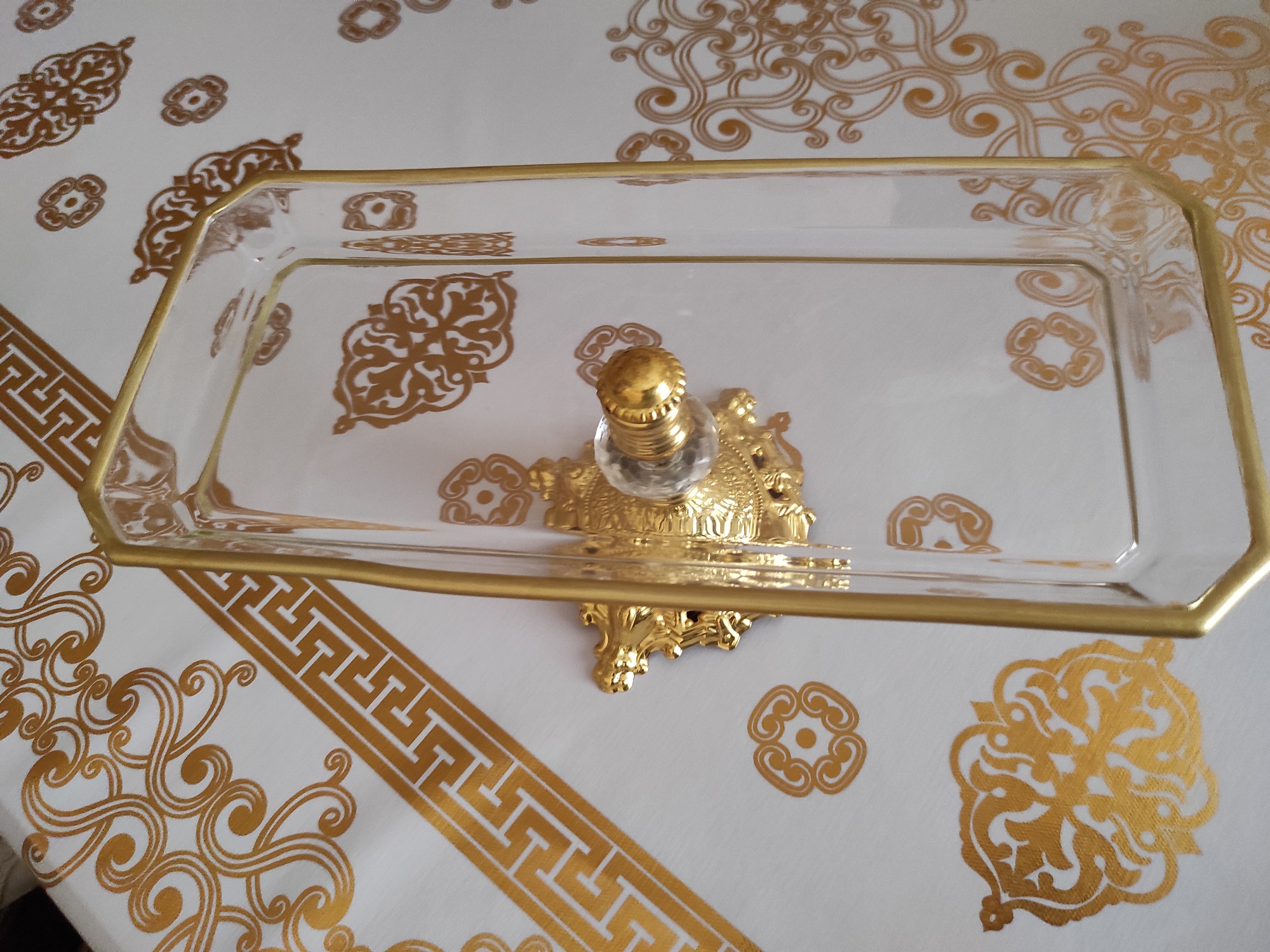The image shows a close-up, elevated view of a rectangular, clear glass tray with a gold rim, placed on a white tablecloth with a gold patterned design. The tablecloth features intricate swirling patterns and a Greek key border. The tray is elevated by a decorative gold-colored stand that is attached to its underside, featuring ornate, filigree-like detailing and a clear, faceted knob on top. The scene appears to be indoors, on a flat surface covered by the tablecloth. There are no people or animals visible in the image. The lighting is bright, casting a slight reflection on the glass tray. The overall aesthetic suggests an elegant or ceremonial setting.