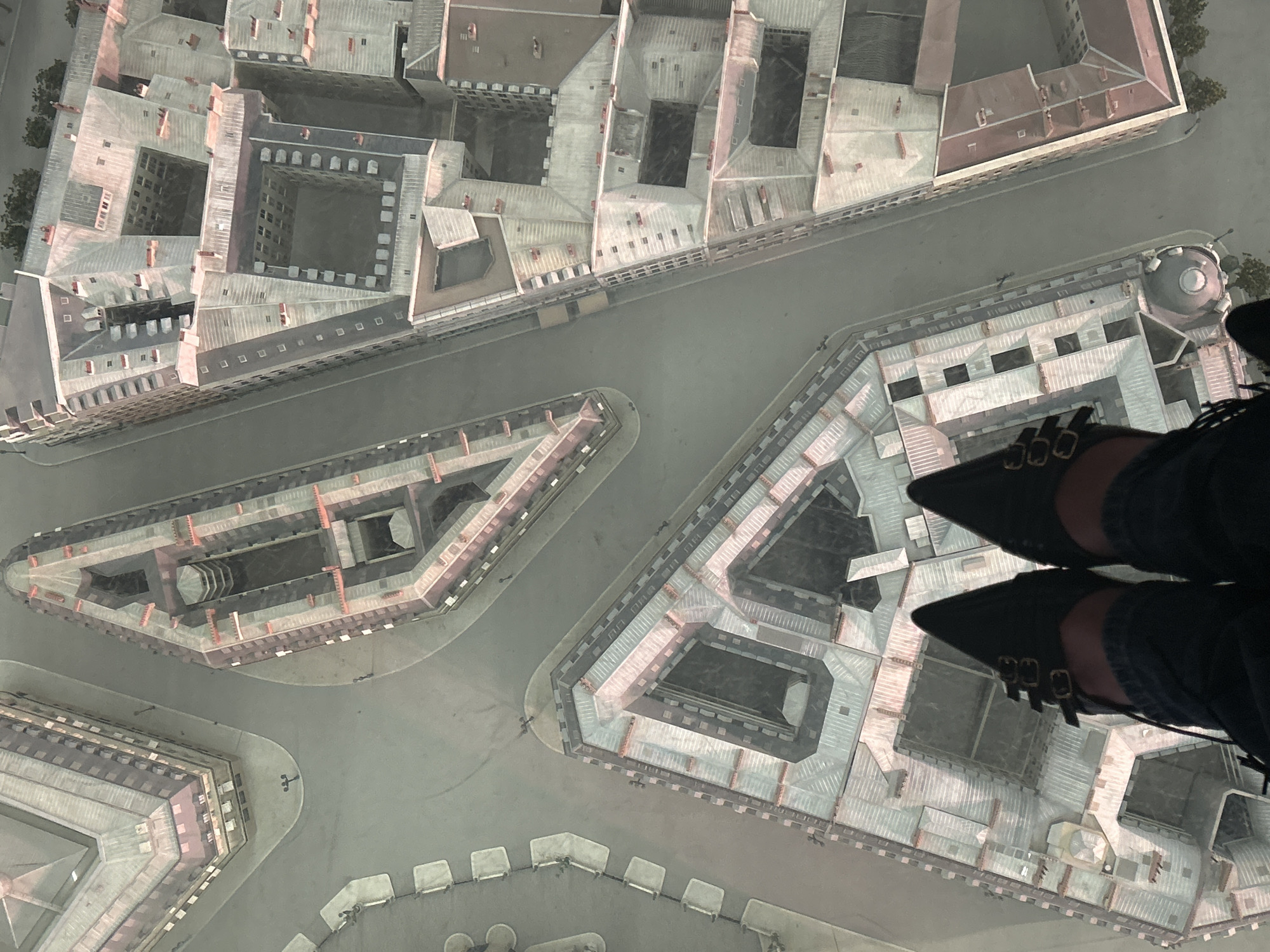 The image presents an overhead perspective of the city of Paris, France, featuring a detailed architectural model of the Place des Vosges, characterized by its symmetric layout of buildings surrounding a central square. The buildings, constructed in a historic style, are primarily light-colored with dark window openings and red roofs. At the bottom right, are a pair of women’s feet with pointed black shoes, wearing jeans. The scene is under flat, artificial illumination, suggesting an indoor setting.