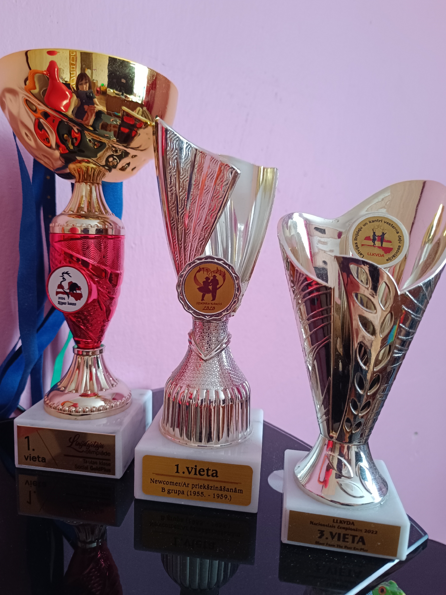 Three trophies are displayed on a dark, reflective surface against a plain light pink wall. Blue ribbons hang behind the leftmost trophy.
The trophy on the left has a gold cup, a red patterned body, and a gold base, resting on a white marble stand. Its plaque reads "1. vieta" (1st place) and "Līnijdejotāju olimpiāde" (Line Dance Olympiad), "Tautas klase" (People's Class), "Social GoldPlus". A circular badge on its body shows "2024 Rīgas kauss" (Riga Cup) with a silhouette of Latvia and a dancer. A reflection of a person, likely the photographer, is visible in the gold cup.
The middle trophy is silver and gold/bronze with a textured body, also on a white marble stand. Its plaque states "1.vieta" (1st place), "Newcomer/Ar priekšzināšanām" (Newcomer/With prior knowledge), "B grupa (1955. - 1959.)". A badge on its body reads "SENTRU KANSS 2020" (Centre Cup 2020) and depicts two dancers.
The trophy on the right is silver and gold/bronze with a decorative, leaf-like pattern, standing on a white marble base. Its plaque indicates "LLKVDA", "NACIONĀLAIS Čempionāts 2022" (National Championship 2022), "3.VIETA" (3rd place), and "Blast From The Past Ex-Plor". A badge near the top reads "Latvijas līnij un kantri vesterna deju asociācija" (Latvian Line and Country Western Dance Association), "LLKVDA", with two dancers.
The trophies represent achievements in line and country western dancing, likely awarded in Tebras, Latvia, as suggested by the language and location context.