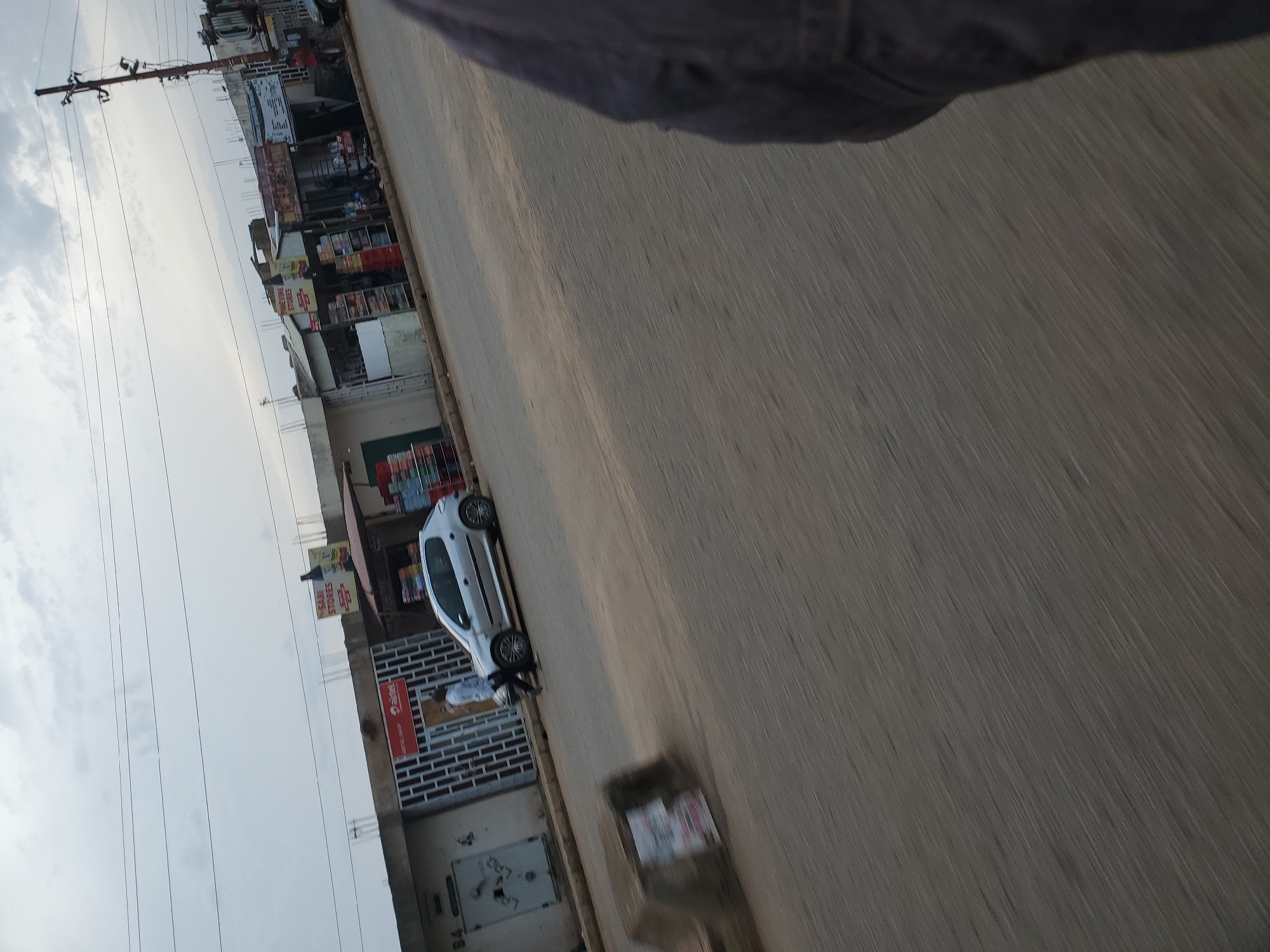 In Bukuru, Nigeria, a street scene unfolds with a silver hatchback car parked on the right side of the road, facing left. A person in a light-colored shirt and dark pants walks in front of the car. To the left of the car are several shops with corrugated metal roofs and open fronts displaying goods. One shop has a sign that reads "AIRTEL SHOP". Further to the left, a building with a closed door and a sign that says "84" is visible. In the foreground, a concrete structure with posters is visible on the left side of the road. The sky above is overcast, and power lines crisscross the frame. The image has a blurred effect, suggesting motion.