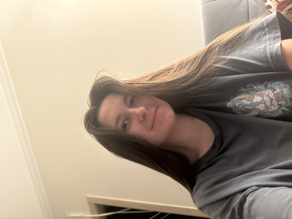 A young woman with long, straight brown hair poses for a selfie. She wears a dark gray T-shirt featuring a graphic design on the chest. The background consists of an off-white wall and a hint of furniture, suggesting an indoor setting. The image is slightly rotated, offering a casual and relaxed perspective. Her expression is soft and seemingly neutral.