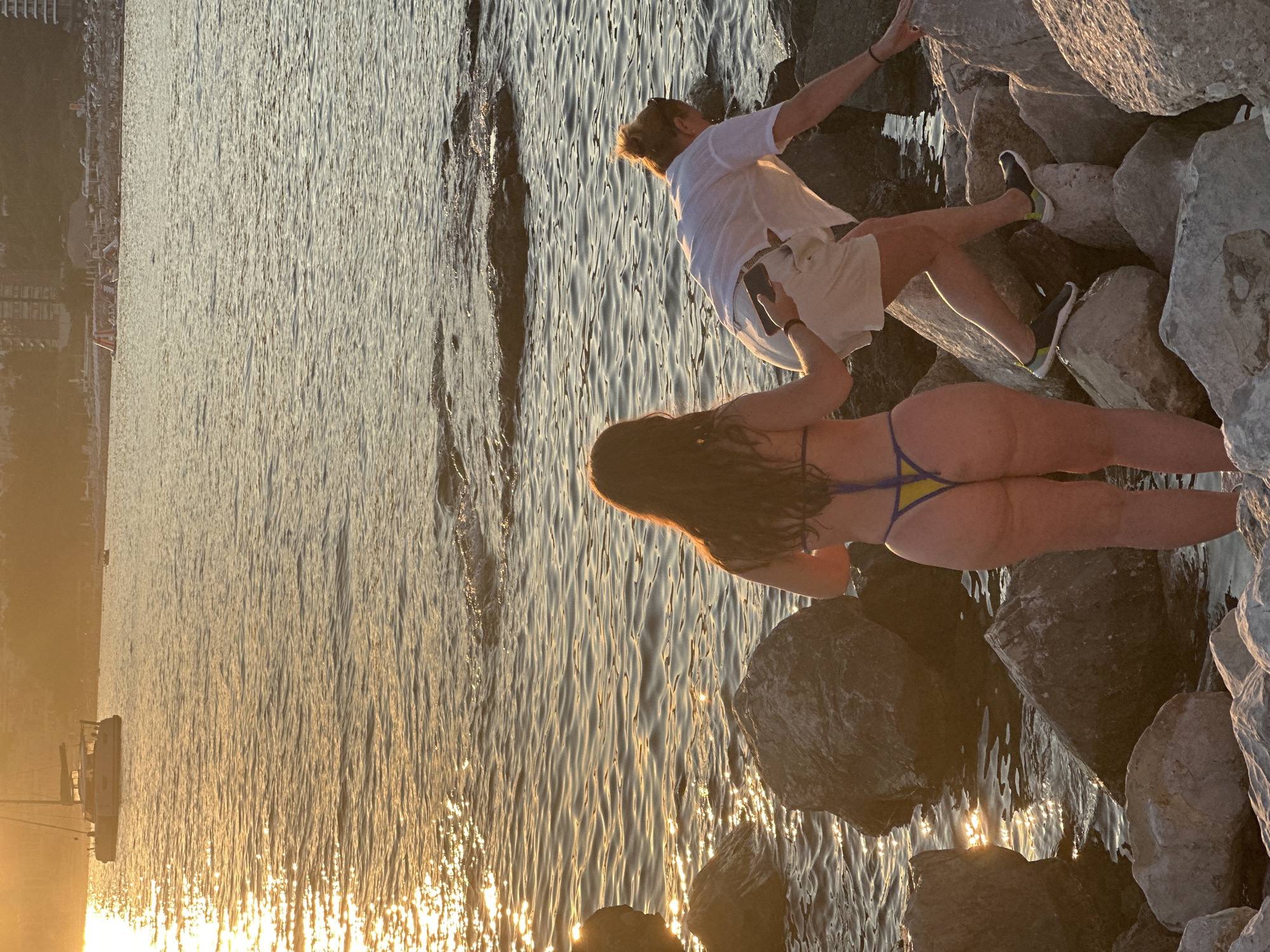 In the city of Rafailovići, Montenegro, two individuals are present on a rocky shoreline as the sun sets. The water is calm with gentle waves lapping against the shore. The light of the setting sun reflects off the water, creating a warm, golden hue. A catamaran is visible in the distance on the water, along with a blurred cityscape in the background. A woman in a bikini stands in the foreground facing away from the camera, while a person in a white shirt and shorts is climbing over the rocks, holding a mobile phone. The rocks are large and uneven, characteristic of a natural coastline. The overall atmosphere is serene, with the natural beauty of the sunset and the coastal landscape dominating the scene.