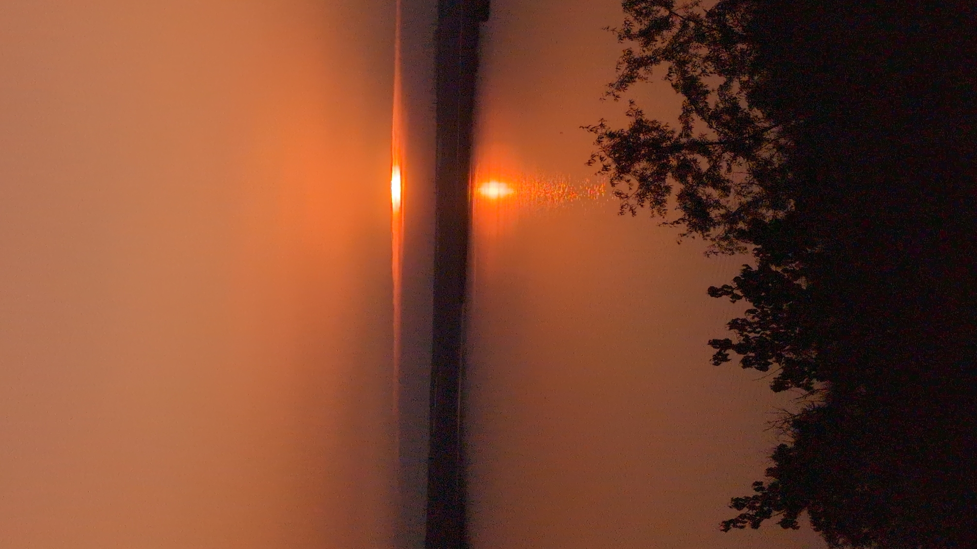 The image shows a sunset over a body of water. The sky is a gradient of orange and red hues, with the sun appearing as a bright, circular orb near the horizon. A thin band of clouds stretches horizontally across the sky, partially obscuring the sun. The sun's reflection shimmers on the water's surface, creating a bright path of light. In the foreground, silhouetted trees with dark foliage are visible, framing the bottom of the scene. The overall atmosphere is calm and serene, indicative of a natural landscape at dusk. The specific location in Kostiantynivka, Ukraine, is not identifiable from the visual elements of the image.