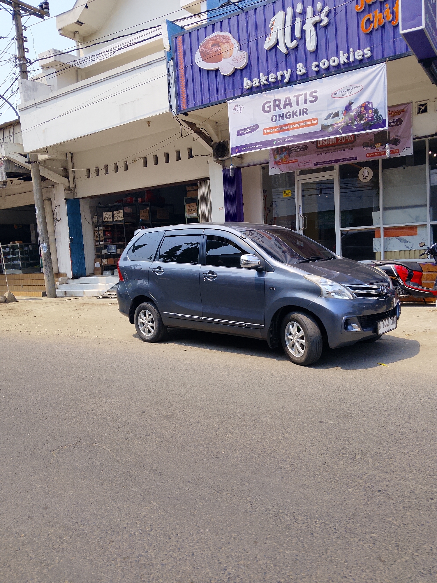 The image depicts a street scene in Ngelo, Indonesia, in front of a bakery and cookie shop named "Alif's Bakery & Cookies". A dark gray minivan, possibly a Toyota Avanza based on its design and the visible emblem, is parked on the right side of the street, facing the viewer. The vehicle's license plate is visible and reads "B 1445 CHV". The shop itself features a prominent purple awning with the bakery's name in white lettering. A large banner is hung across the storefront advertising "GRATIS ONGKIR" (free delivery) with a slogan stating "tanpa minimal jarak/radius km" (without minimum distance/radius km). Another banner below advertises "DISKON 20%" (20% discount). To the right of the minivan, a red and black motorcycle is partially visible, parked near the shop entrance. The street in front of the building is paved asphalt, and a sidewalk is present. The building itself is white with large glass windows and doors. The sky above is bright and appears to be daytime.
