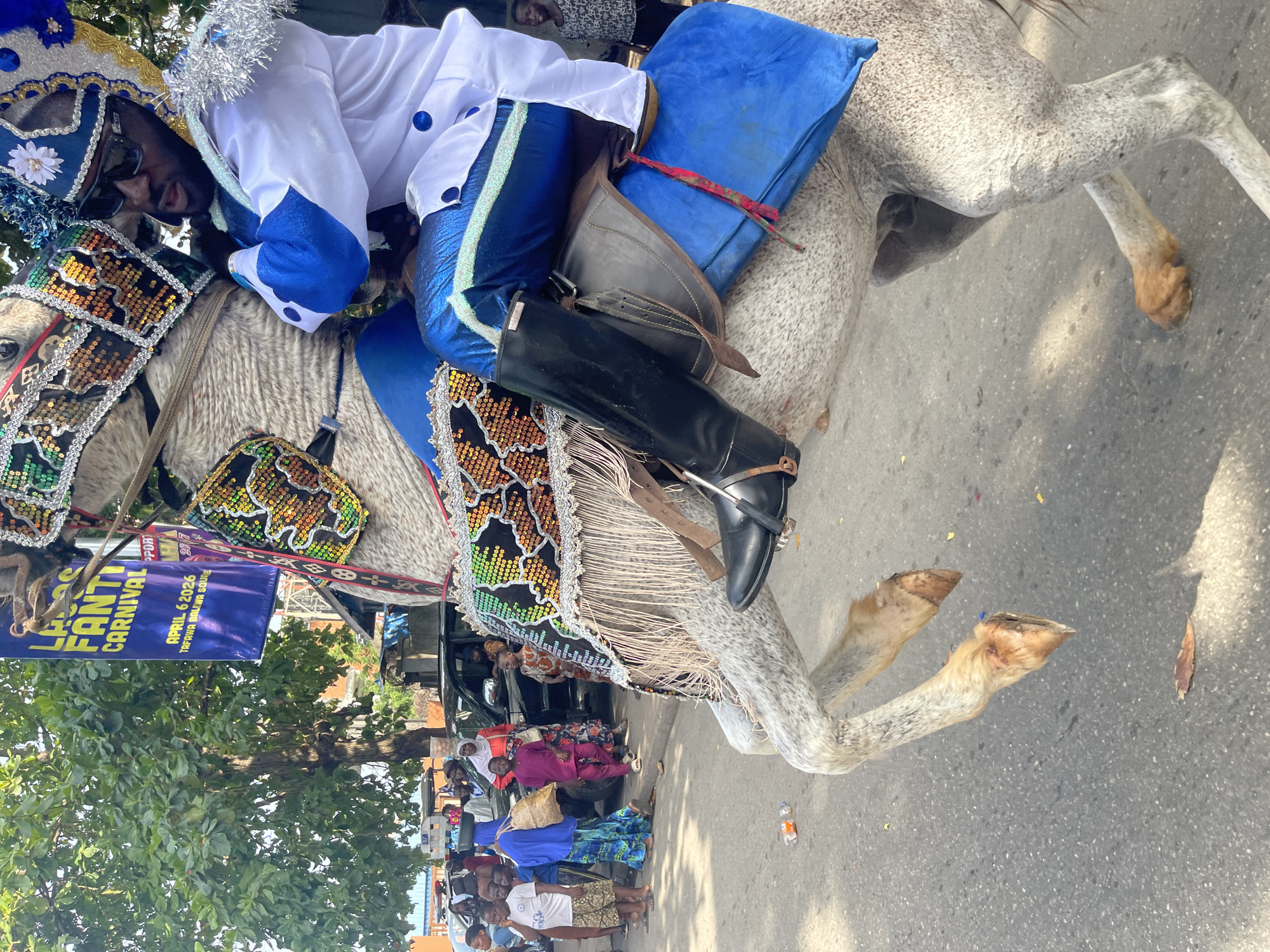 A man in a white and blue costume rides a decorated gray horse on a street in Lagos, Nigeria, as spectators watch. The banner reads "LAGOS FANTI CARNIVAL APRIL 6, 2026, TAFRWA BALEWA SQUARE".