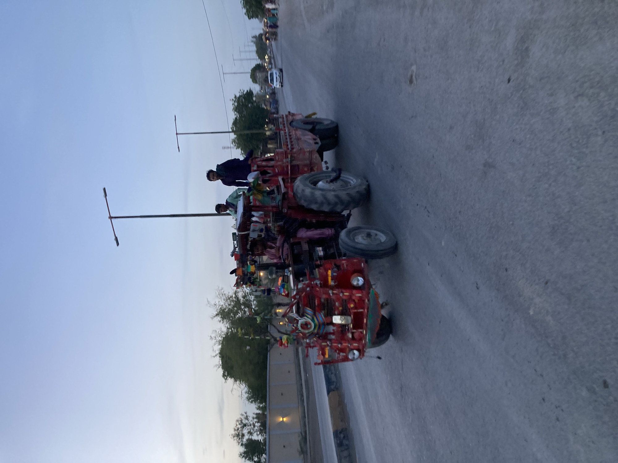 The image captures a vibrant, heavily decorated red tractor or power tiller pulling a loaded trolley along an asphalt road at dusk in Uthal, Pakistan. The vehicle, adorned with numerous colorful ribbons, streamers, and small decorative items, suggests a personalized or possibly festive appearance.
Two men are prominently visible riding on the loaded trolley. One man, dressed in a dark shirt, stands near the front of the trolley, while another man, wearing a lighter green or blue shirt, is positioned behind him, either standing or sitting among the cargo. The trolley itself is laden with various items, possibly covered by tarpaulins or fabrics, indicating its use for transporting goods.
The setting is a paved road, which appears to be well-used. Alongside the road, there are trees with green foliage and streetlights, some of which are simple poles. In the distance, a white car is visible traveling in the opposite direction on the same road. The sky is a clear, light blue, suggesting the time of day is either early evening or dawn, with soft, diffused lighting across the scene. The overall atmosphere appears peaceful and typical of daily life. No discernible text is visible on the vehicle or in the surroundings.