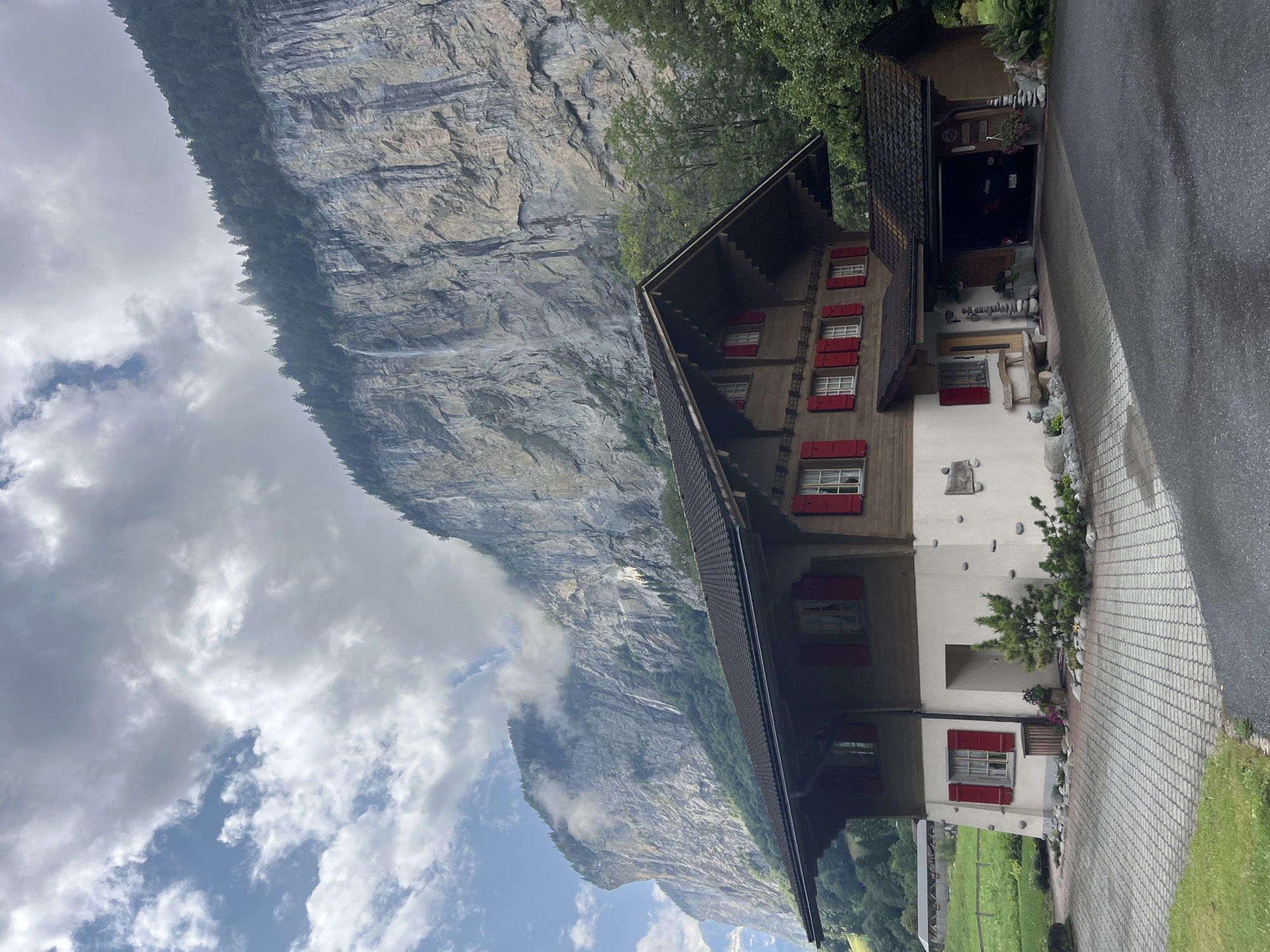 The image shows a two-story wooden house with red shutters, situated in front of a steep, rocky cliff face with waterfalls cascading down. The house has a dark tiled roof with wide eaves. The lower level is partially rendered and decorated with decorative stones. There is a paved driveway in front of the house, leading to a garage attached to the right side. A wooden bench is placed outside the front door. Lush green trees and vegetation surround the property, and a meadow with a fence is visible to the left. The sky is partly cloudy, with dramatic cloud formations. The scene is characteristic of Wengen, Switzerland, a village known for its dramatic alpine scenery. No people or animals are visible in the image.