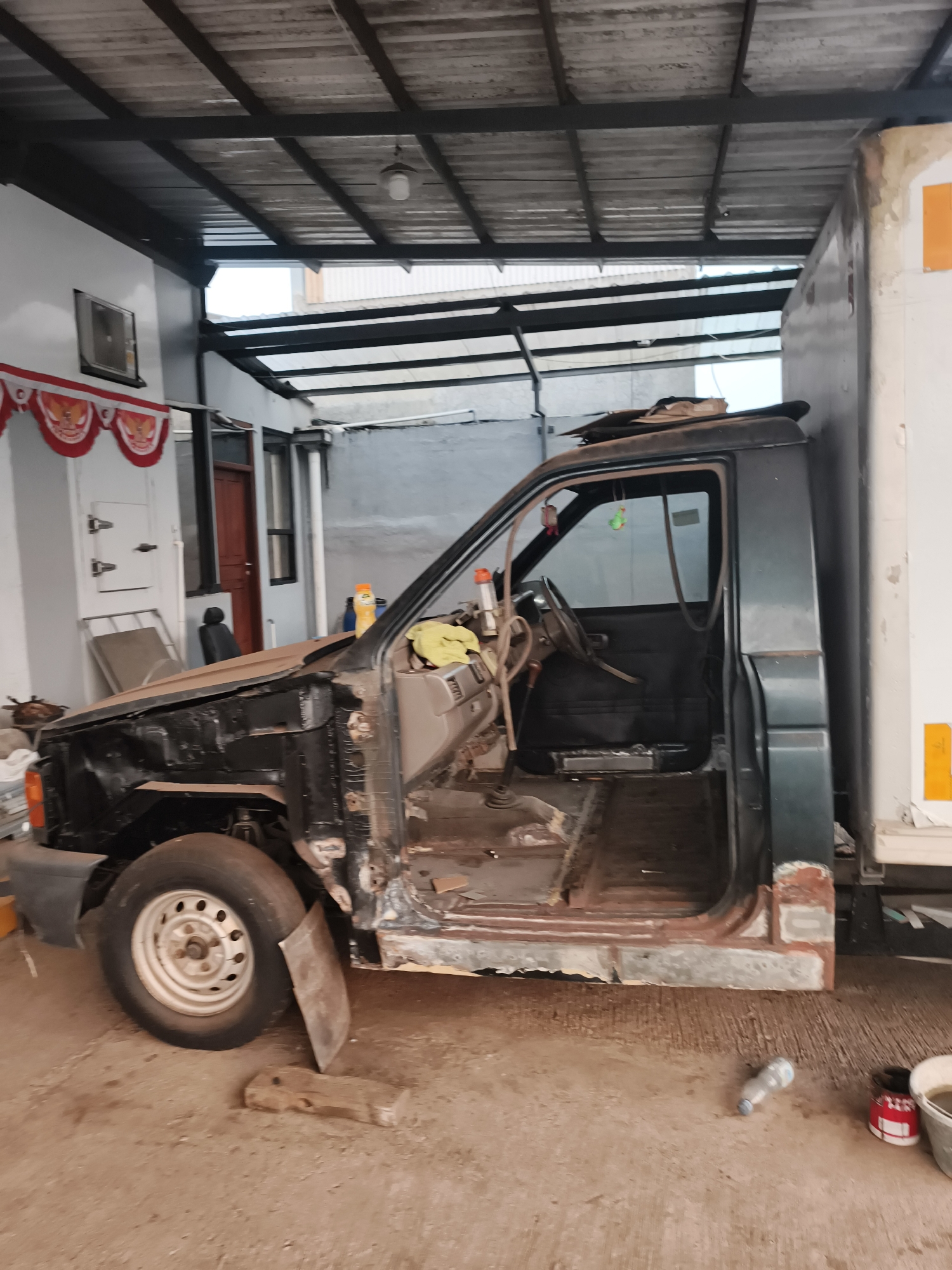 The image shows a dismantled pickup truck in what appears to be a garage or workshop setting, possibly located in Pekalongan, Indonesia, based on the context provided. The truck is a dark green pickup with its hood, doors, and some interior components removed, revealing the frame and chassis. Rust is visible on the exterior of the truck, particularly around the wheel wells and door frames, indicating it is likely undergoing restoration or is in a state of disrepair. The vehicle is positioned next to a large white truck or box body, which appears to be parked closely.
The environment consists of a concrete floor with some scattered debris, including pieces of wood and a plastic bottle. Above the truck, a corrugated metal roof supported by dark metal beams is visible. To the left of the truck, there is a white wall with a small window and red and white decorative banners hanging from the eaves. A brown wooden door is also visible on the left side. The lighting suggests it is daytime, but the interior is somewhat dimly lit. There are no people visible in the image, and the focus is solely on the condition and setting of the pickup truck.