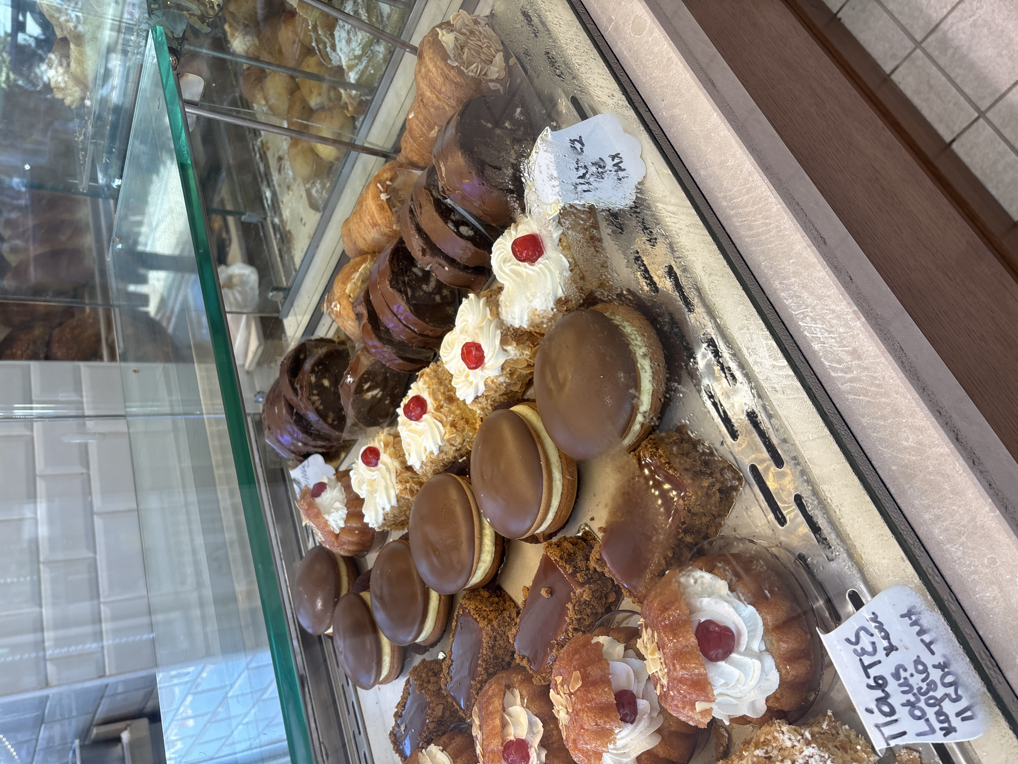 The image shows a display case filled with various pastries, likely in a bakery in Nea Kallikrateia, Greece. The pastries include chocolate-covered cakes, cream-filled buns topped with cherries, and other sweet treats. A handwritten price tag in Greek is visible in the foreground. The display case is made of glass and metal, and the background shows tiled walls and other bakery items. The lighting suggests an indoor environment, possibly a shop. There are no people visible in the image.