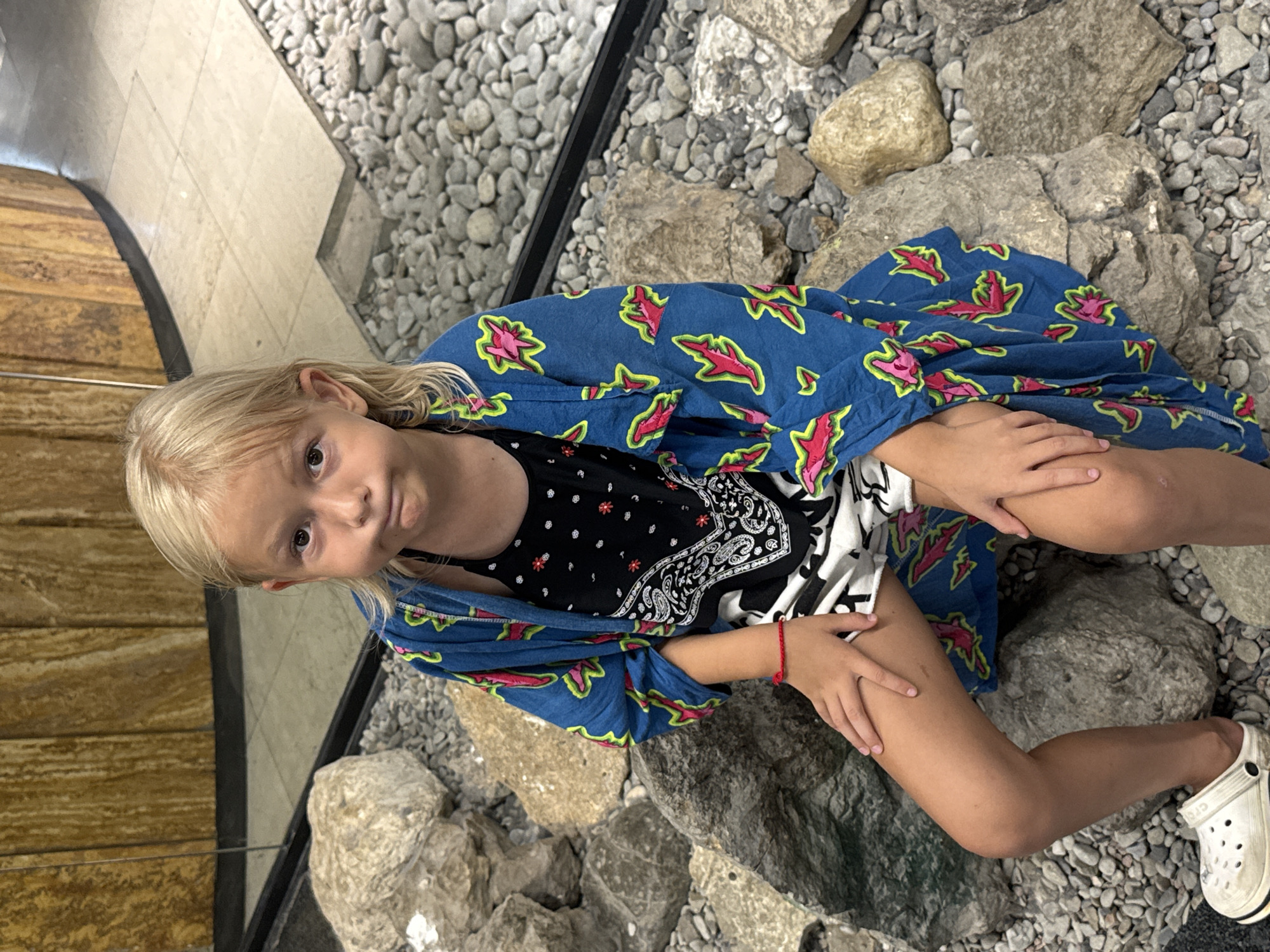 This is a vertical, eye-level, full shot of a young girl sitting on a rocky surface. The girl has blonde hair and is looking at the camera with a neutral expression. She is wearing a black top with a white bandana-like pattern and a blue open robe with a pink and green leaf-like pattern. Her left hand rests on her knee, and her right hand is on her upper thigh. She has a red string bracelet on her left wrist. She is wearing a white clog on her right foot. The background consists of numerous grey and beige rocks and pebbles. To the left of the rocks is a wooden surface and a black border. The scene appears to be outdoors, possibly in a decorative garden or walkway. The lighting suggests it is daytime. The location is identified as Yalta, Ukraine.