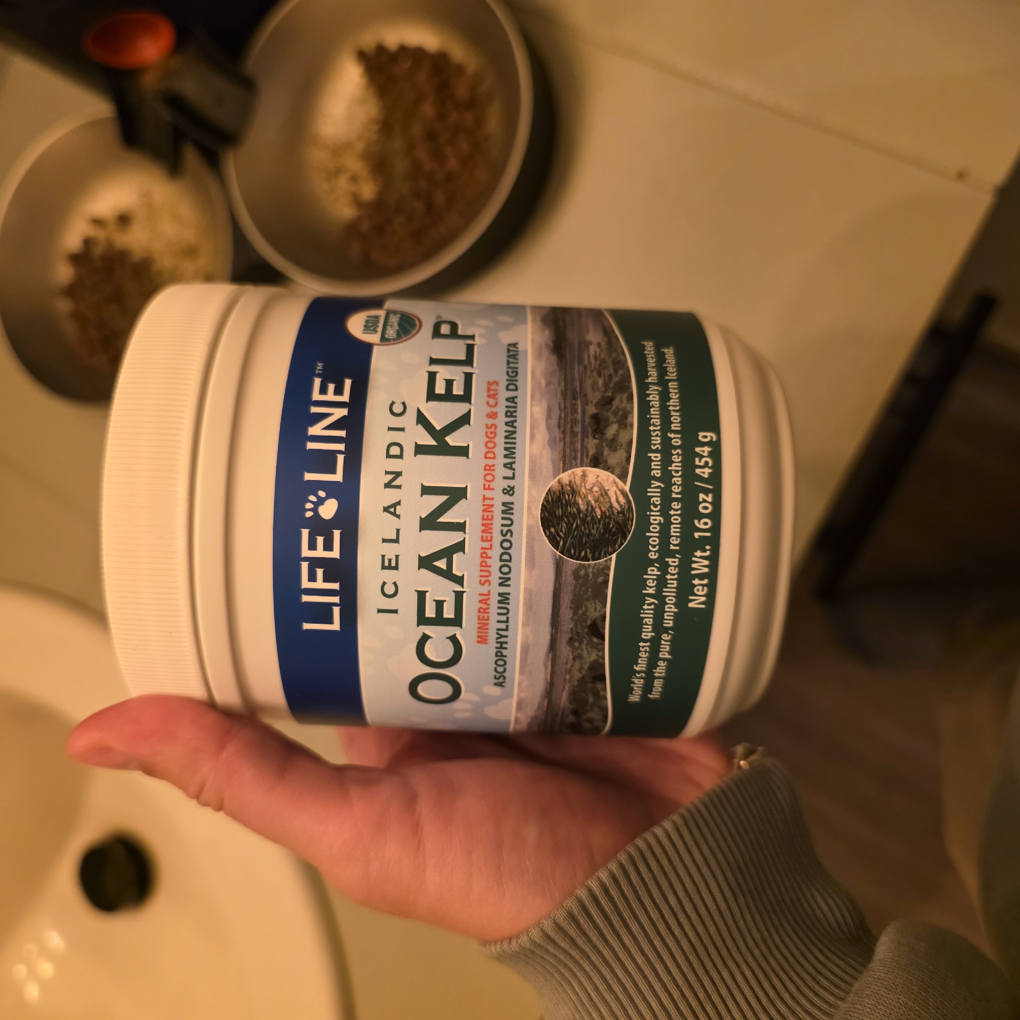 A person's hand holds a white plastic container labeled "LIFE LINE ICELANDIC OCEAN KELP MINERAL SUPPLEMENT FOR DOGS & CATS". The container is partially obscuring a bathroom sink with a black drain in the background. Two metal bowls containing brown kibble are visible to the right of the container. The image appears to have been taken in a home bathroom. The provided context about the city of Beaverton, United States of America, is not visually verifiable from the image itself.