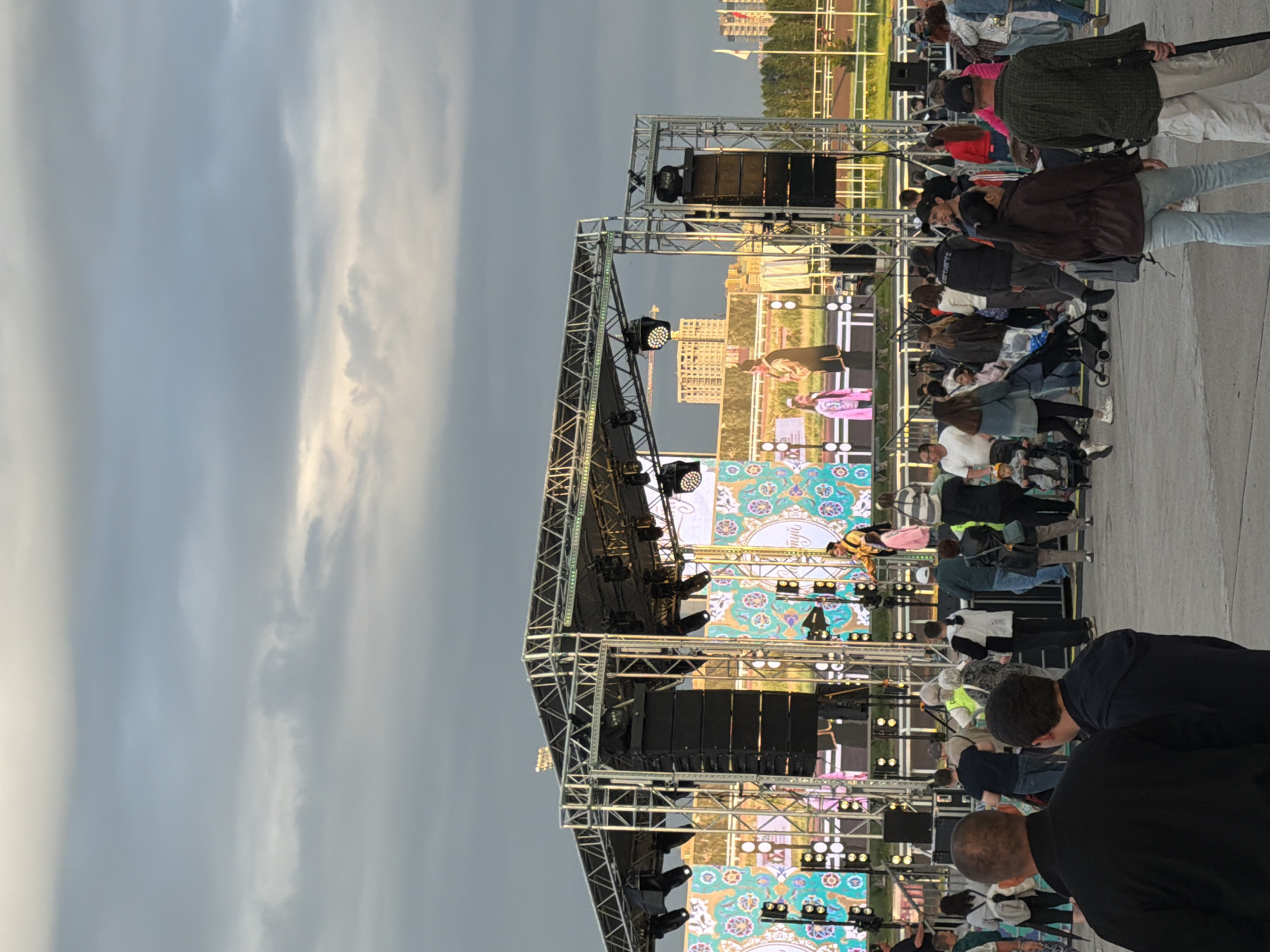 The media file shows an outdoor event with a stage set up for a performance. The stage is constructed from metal scaffolding and features multiple rows of speakers and lighting equipment. A large screen behind the stage displays a colorful, patterned backdrop, and on it, figures in traditional clothing appear to be performing or being displayed. The scene is viewed from ground level, looking up towards the stage.
A crowd of people is gathered in front of the stage, facing it. They are mostly standing, with some individuals sitting or pushing strollers. The people are dressed in casual, everyday clothing, suggesting a relaxed atmosphere. Some are looking towards the stage, while others are interacting with each other.
The time of day appears to be late afternoon or early evening, as indicated by the lighting and the sky. The sky is overcast with some lighter patches where the sun might be breaking through or reflecting off clouds. The overall setting seems to be an open public space, possibly a park or a plaza, given the paved ground and the distant buildings visible behind the stage.
There are no discernible emotions visible in the crowd due to the distance and resolution. The activity taking place is clearly a public performance or entertainment event. The backdrop on the screen features intricate patterns that suggest a cultural or festive theme. The overall impression is one of a community gathering for an outdoor concert or show.
