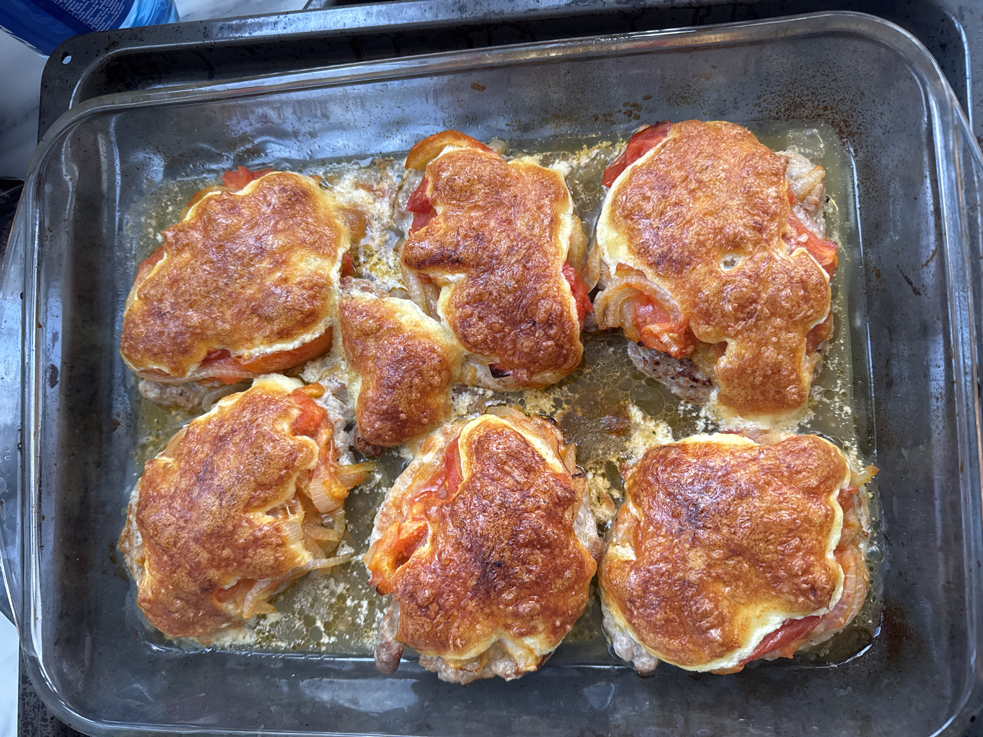 This media file shows a top-down view of a glass baking dish filled with six golden-brown, baked chicken cutlets. Each cutlet is topped with sliced tomatoes and onions, all covered with a melted, bubbly cheese crust. The cheese is browned and slightly crispy in places, with some areas showing a golden-brown hue. The chicken cutlets are arranged in two rows of three within the rectangular glass dish, which appears to be sitting on a countertop. The surrounding area is out of focus, but a portion of a dark metal baking tray is visible around the glass dish. The overall scene suggests a home-cooked meal, possibly after being taken out of an oven. The lighting appears to be artificial, suggesting the photo was taken indoors. There is no visible text in the image. The setting is a kitchen, likely in Sochi, Russia, given the provided context. The food appears to be freshly baked and ready to be served.