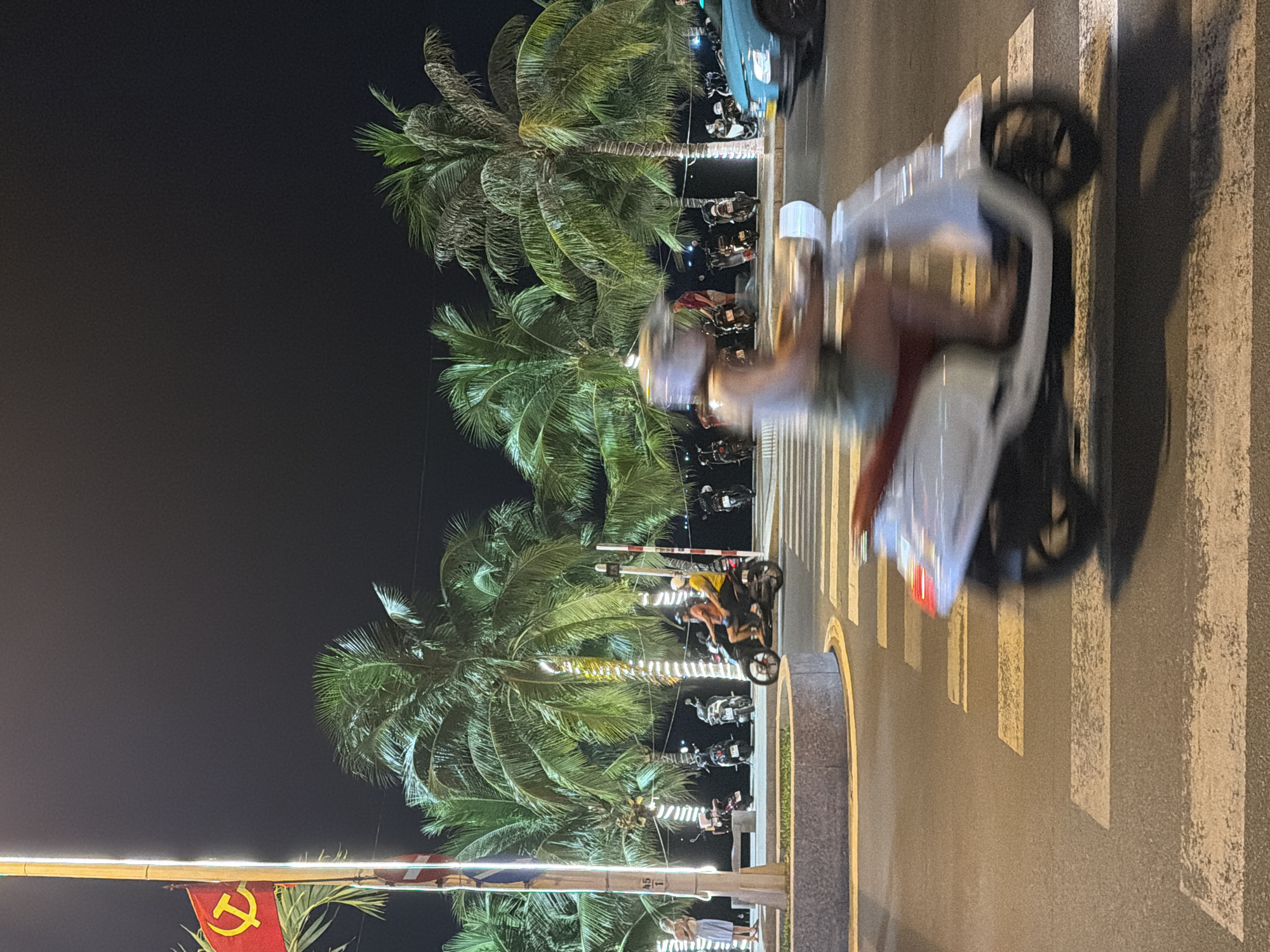 The image depicts a night scene on a street in Nha Trang, Vietnam, characterized by palm trees and illuminated by streetlights and decorative lighting. In the foreground, a person on a white scooter is captured in motion, crossing a pedestrian crossing. The scooter's wheels and the rider are blurred due to the movement. Behind the scooter, another motorcycle with two individuals, one wearing an orange shirt and the other a yellow shirt, is also moving. Several other motorcycles are parked along the sidewalk to the left and right. A flag, likely the Vietnamese national flag with a yellow star on a red background, is visible on a pole to the far left, partially obscured by a palm tree decorated with string lights. A woman in a light-colored dress stands on the sidewalk to the left, looking at her phone. The environment suggests a tropical coastal city at night, with the presence of palm trees and a well-lit street, indicative of a public promenade or a busy thoroughfare.
