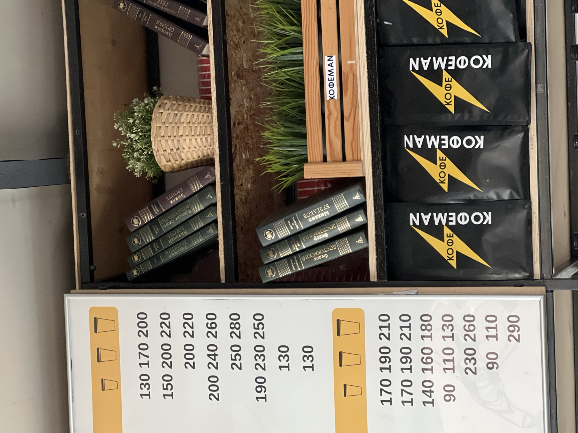 This image shows a shelf unit in what appears to be a café or store, possibly in Berezniki, Russia. The shelves hold a variety of items including:
* Black and yellow branded coffee packets labeled "KOΦΕΜΑΝ" (KOFE in Cyrillic).
* Several stacks of books, with spines showing titles in Russian: works by authors such as Mikhail Bulgakov, Fyodor Dostoevsky, Emily Bronte, Nikolai Gogol, F. Scott Fitzgerald, and George Orwell
* Decorative green plants, including one in a woven basket.
* A price list for coffee drinks with prices in Russian rubles.
The presence of Russian text on the books and price list suggests the location is in Russia, potentially Berezniki if that is the actual location.