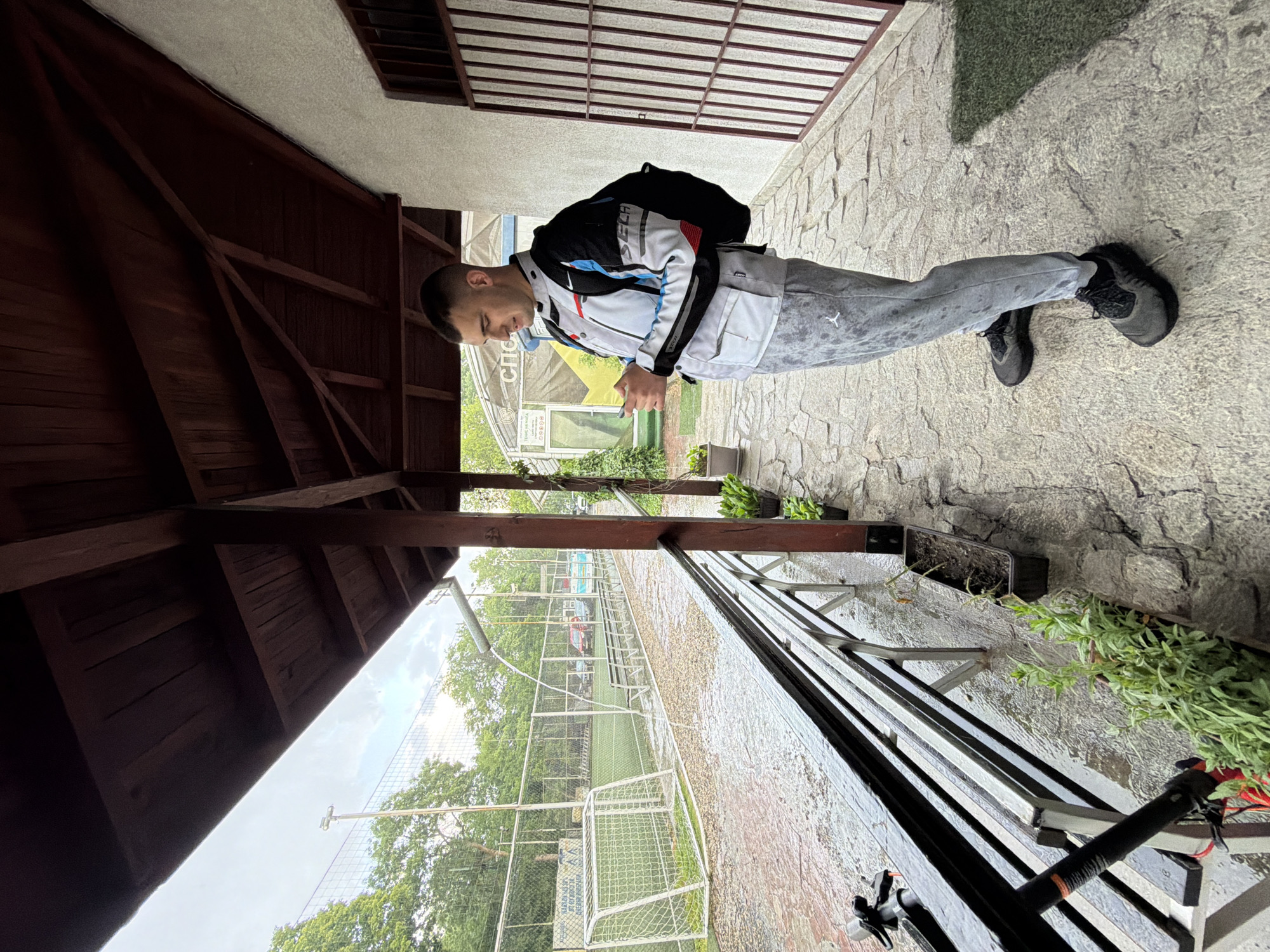 A young man stands in an alleyway in Sofia, Bulgaria. He is wearing grey sweatpants with a white logo, a white, blue, red, and black jacket, and dark shoes. A black backpack is on his back. He looks down at an object in his hands, possibly a phone. The alleyway is paved with cobblestones. One side of the alley is a wooden structure, possibly a covered seating area. The other side has a view of a soccer field with a net, through a metal fence. The weather appears cloudy. The location is assumed due to the soccer field often found in Sofia.
