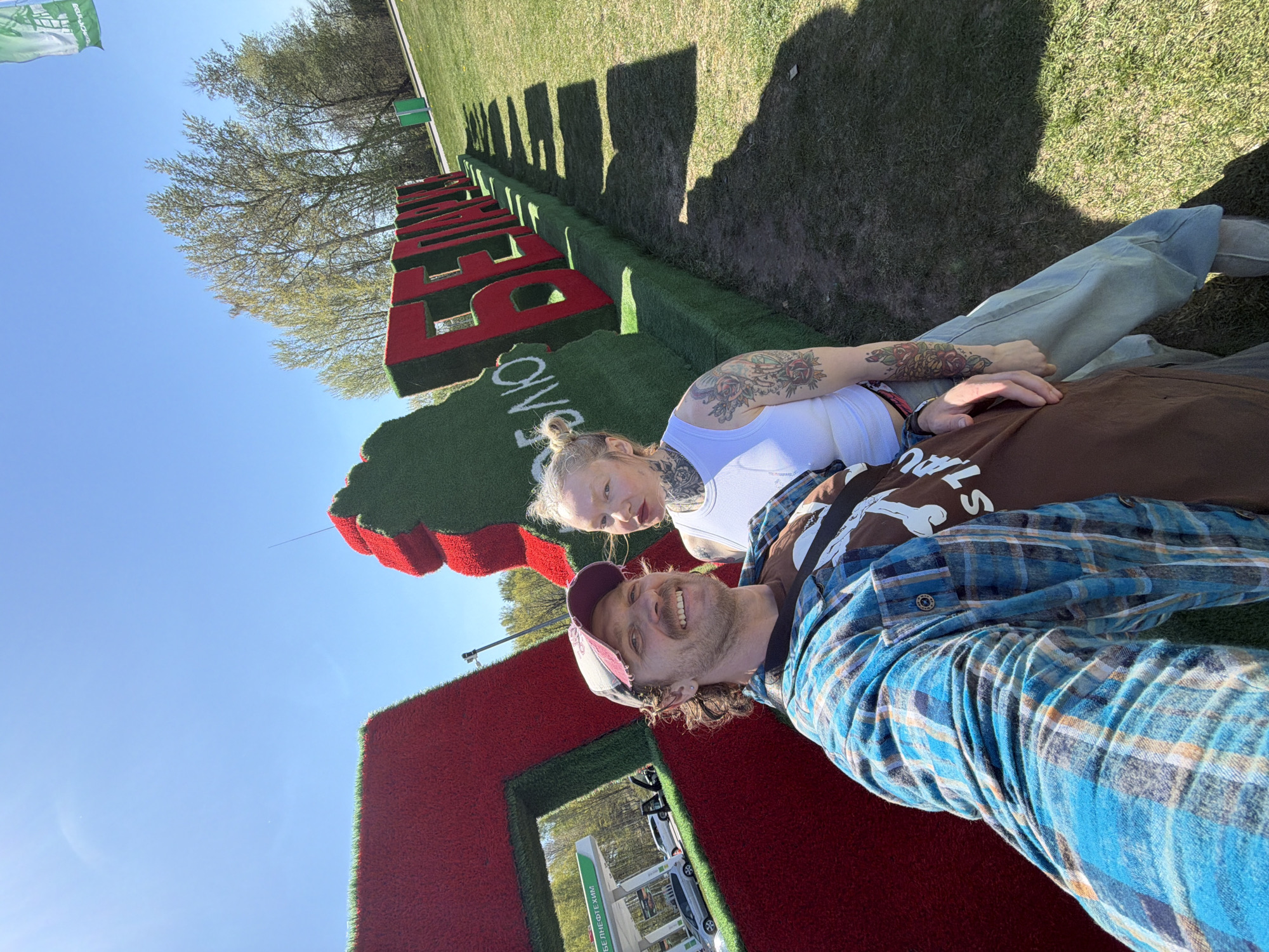 Two people are lying on a large, grass-covered structure that spells out "BELARUS". The structure is made of red and green grass and is set on a grassy field under a clear blue sky. One person, presumably a man, wears a blue plaid shirt and a burgundy baseball cap, smiling at the camera. The other person, presumably a woman, wears a white tank top and has visible tattoos on their arms and neck. They are both lying down, possibly taking a selfie or having someone else take the photo. In the background, there are trees and a gas station. The location is Крошынскі сельскі Савет, Belarus.