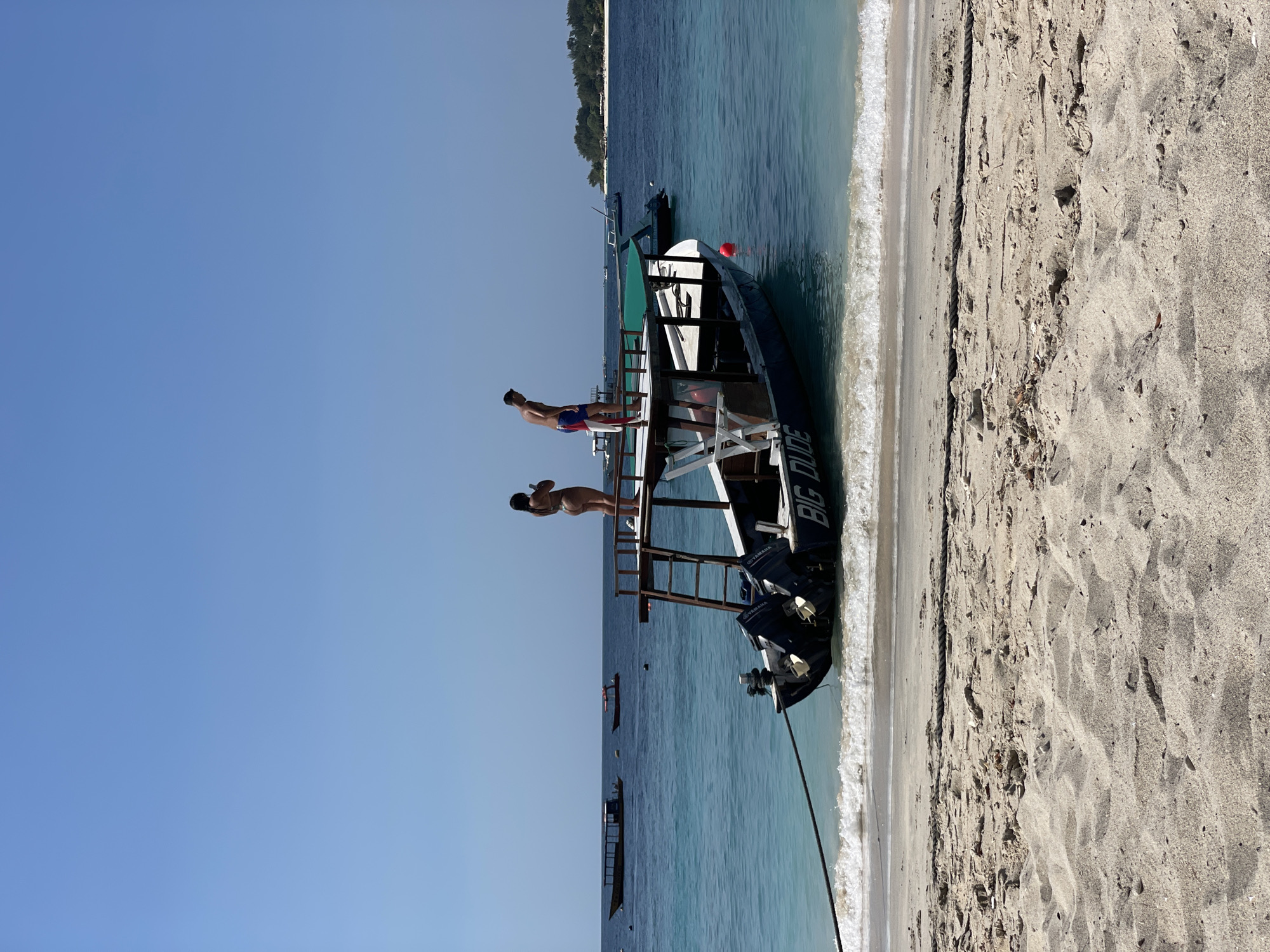 A wide shot captures a clear, sunny day at a tropical beach. In the foreground, the sandy beach slopes gently towards the turquoise water, with small waves lapping at the shore. A thick rope lies on the sand, extending into the water.
Anchored close to the shore is a long, dark-hulled boat with a raised wooden platform at the stern. Two outboard motors are mounted on the back of the boat. The words "BIG DUDE" are visible in white lettering on the side of the hull.
On the wooden platform of the boat, two individuals are present. A woman in a bikini stands on the left, holding a mobile phone. A man in swim trunks stands to her right, facing away from the camera and looking out at the water.
In the background, the clear blue ocean stretches out, dotted with other boats of various sizes. Further in the distance, a lush green island with a white sandy beach is visible under the bright blue sky. The horizon is sharp and clear, indicating good visibility.