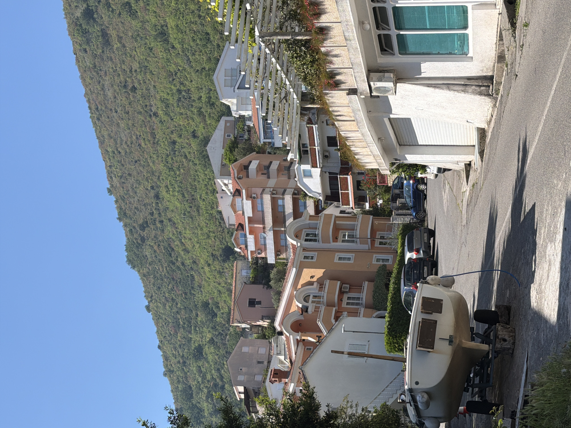 The image captures a sunny, clear day in Petrovac centar, Montenegro, depicting a residential street flanked by multi-story buildings and a steep, wooded mountain. The perspective is from slightly above the street, looking down its length.
In the immediate foreground on the right, a light-colored boat rests on a trailer, parked alongside the road. The boat's stern, featuring two dark, rectangular hatches, is visible. A blue hose extends from near the boat onto the asphalt. Green foliage partially obscures the bottom right.
The street, made of asphalt, extends into the mid-ground. Strong, elongated shadows from unseen trees or structures are cast across its surface, suggesting it is either morning or late afternoon. Cars are parked along the right side of the road, including a blue hatchback and a silver or grey vehicle.
To the right of the street, a series of multi-story buildings, likely apartments or homes, ascend a gentle slope. These structures feature varying shades of white, beige, and light orange, with many sporting red-tiled roofs and balconies. Several air conditioning units are affixed to their exteriors. A prominent white building in the mid-right has large windows with teal or blue blinds/shutters and a white garage door beneath. Higher up, a white pergola adorned with greenery is visible.
Dominating the left side of the frame is a large, very steep mountain face, densely covered in vibrant green trees and vegetation, extending from the mid-ground to the distant background. Above the mountain and between the buildings, the sky is a vivid, cloudless blue.
No people are visible in the scene, and no discernible text is present on any signs, vehicles, or buildings. The overall atmosphere is tranquil and residential, characteristic of a coastal town.