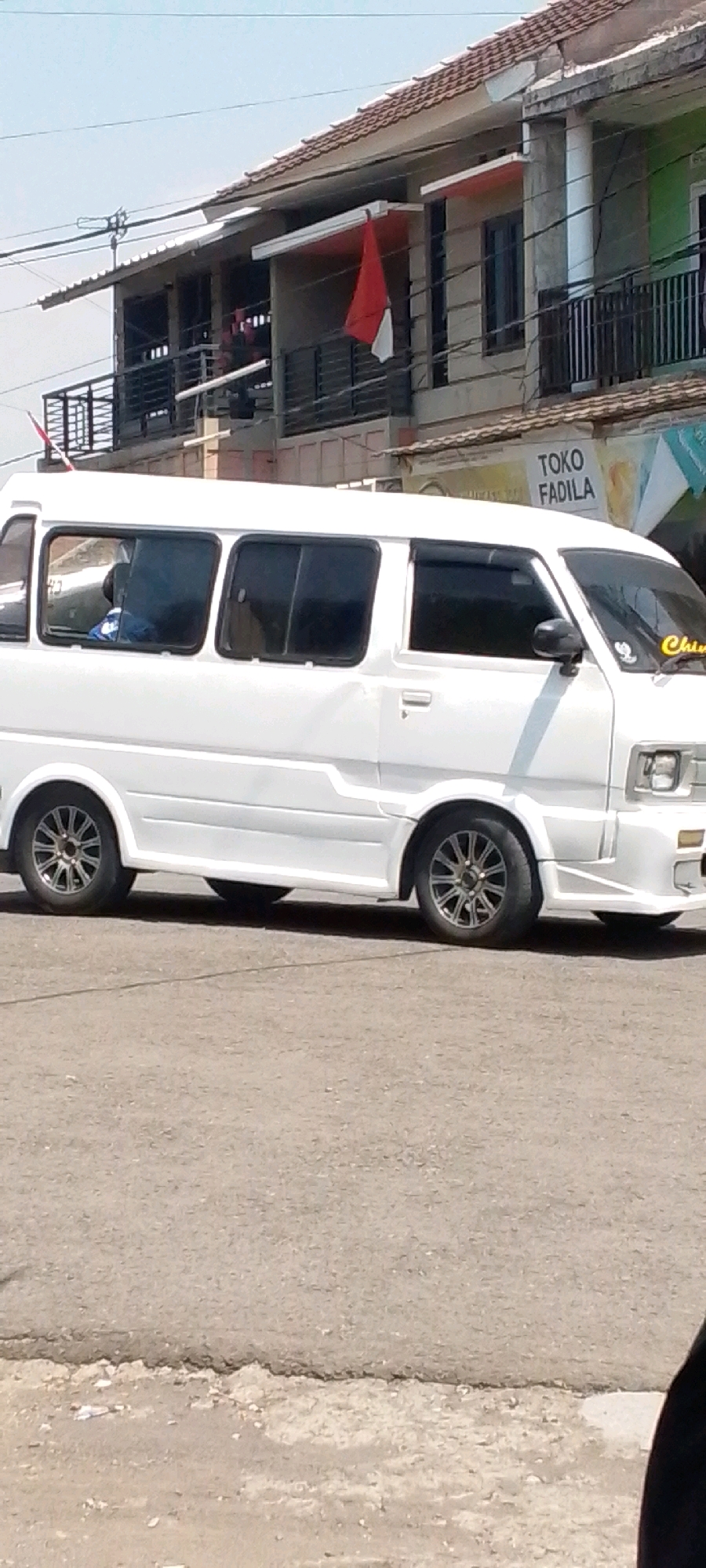 In Cilaku, Indonesia, a white van is parked on a paved street, facing right. The van has tinted windows and a body kit. On the van's windshield, a yellow sticker with the word "Chiri" is visible. Behind the van, a building with a tiled roof stands. The building has a balcony with black railings and two red and white Indonesian flags displayed. A sign on the building reads "TOKO FADILA." The sky is clear and blue.