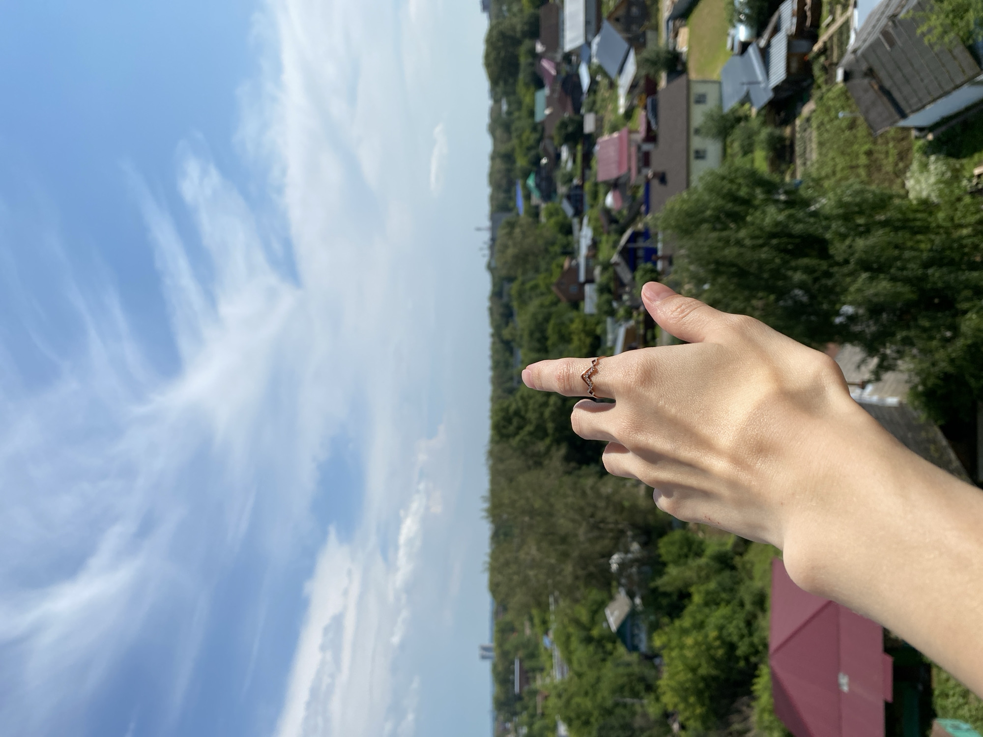 In this medium shot, a person's left hand, adorned with a delicate ring on the index finger, is raised, pointing towards a bright, partly cloudy sky. The sky is filled with wispy white clouds against a clear blue backdrop. Below the sky, a sprawling, tree-filled suburban landscape unfolds, characteristic of the outskirts of Glazov, Russia. The view encompasses numerous houses with varied roof colors, including prominent red-tiled roofs, interspersed with lush green trees. Some houses appear to be single-story dwellings with gardens, while others are larger structures. In the distance, faint outlines of industrial buildings or taller structures can be discerned, hinting at the proximity of more developed areas within the city. The overall scene depicts a sunny day in a residential area, with the raised hand suggesting observation or a gesture directed at the sky.