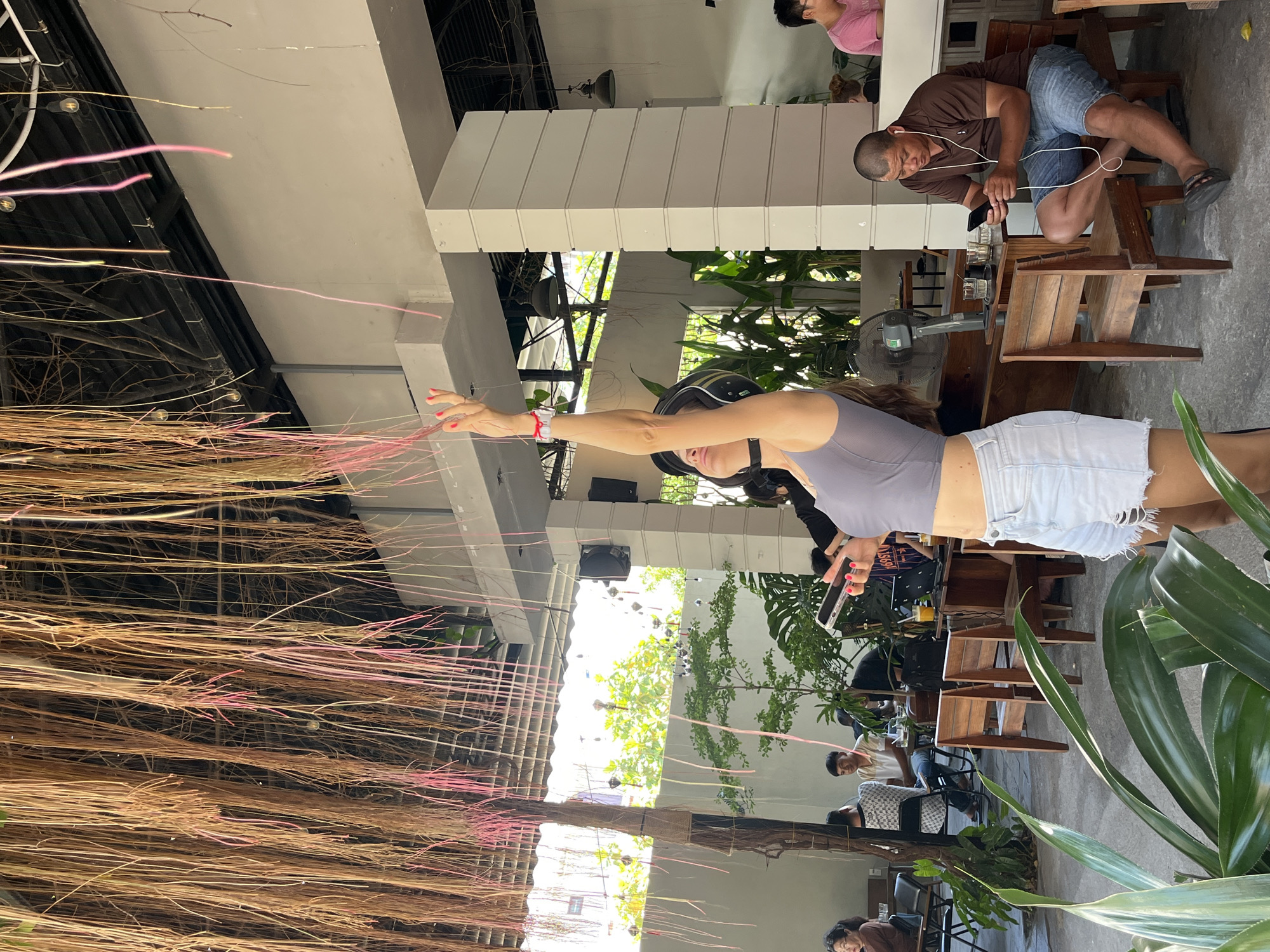 The image depicts an outdoor seating area of what appears to be a cafe or restaurant, likely in Bac Nha Trang Ward, Vietnam. A woman wearing a motorcycle helmet, a gray top, and denim shorts stands with her arm extended. The cafe's interior is partially visible with customers seated at wooden tables and chairs. Man in brown shirt using smartphone. The environment has lush green plants, and an overhead decoration made of light-colored wooden slats from which hang brown and pink colored strands.