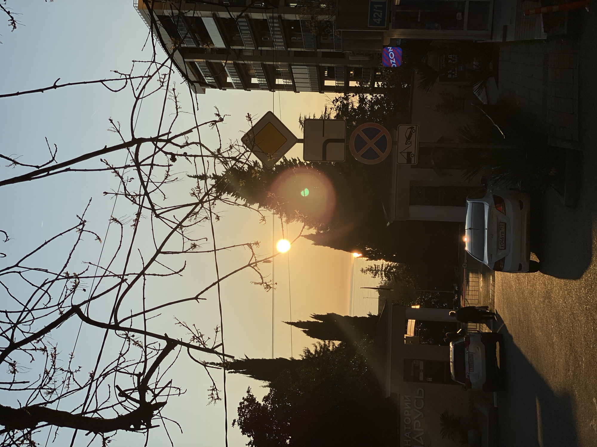 The image shows a street scene in Sochi, Russia, captured during sunset or sunrise. In the foreground, bare tree branches are silhouetted against the sky. A building stands at the right angle, with a sign reading "42/1". Road signs are visible, including a yellow diamond-shaped sign indicating the main road, a sign showing a right turn, and a sign with a blue circle and a red "X" indicating a no-parking zone. The street features parked cars and at least one person in the shade. The car in the foreground has license plate number "k046yb 193". The name “НАТОРРИЙ АРУС” is also visible on a building. The sun is low on the horizon, creating lens flare and casting long shadows along the street. The sky is a gradient of colors from blue to orange. The sea is in the background on the horizon.