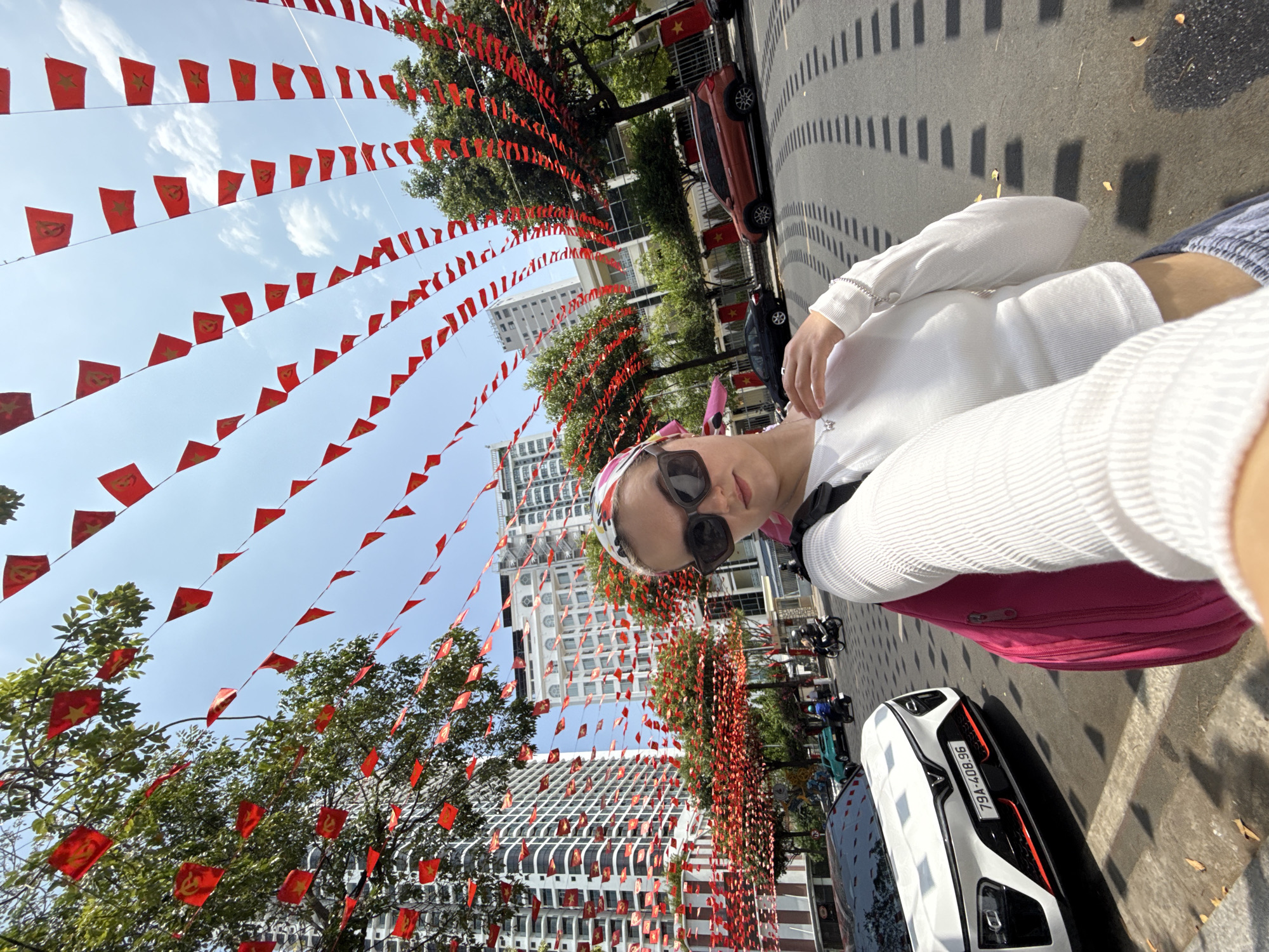 A selfie-style, medium-shot photograph shows a woman on a street decorated with red flags. The location is Nha Trang, Vietnam. The woman is in the foreground, wearing a white top, sunglasses, and a patterned head covering. A pink bag is also visible. The street scene includes cars, scooters, and trees. Red flags with yellow stars are strung above the street. High-rise buildings are visible in the background. The license plate of a white car reads "79A-408.96." The lighting suggests daytime, with a clear sky. Shadows from the trees and decorations mark the street.