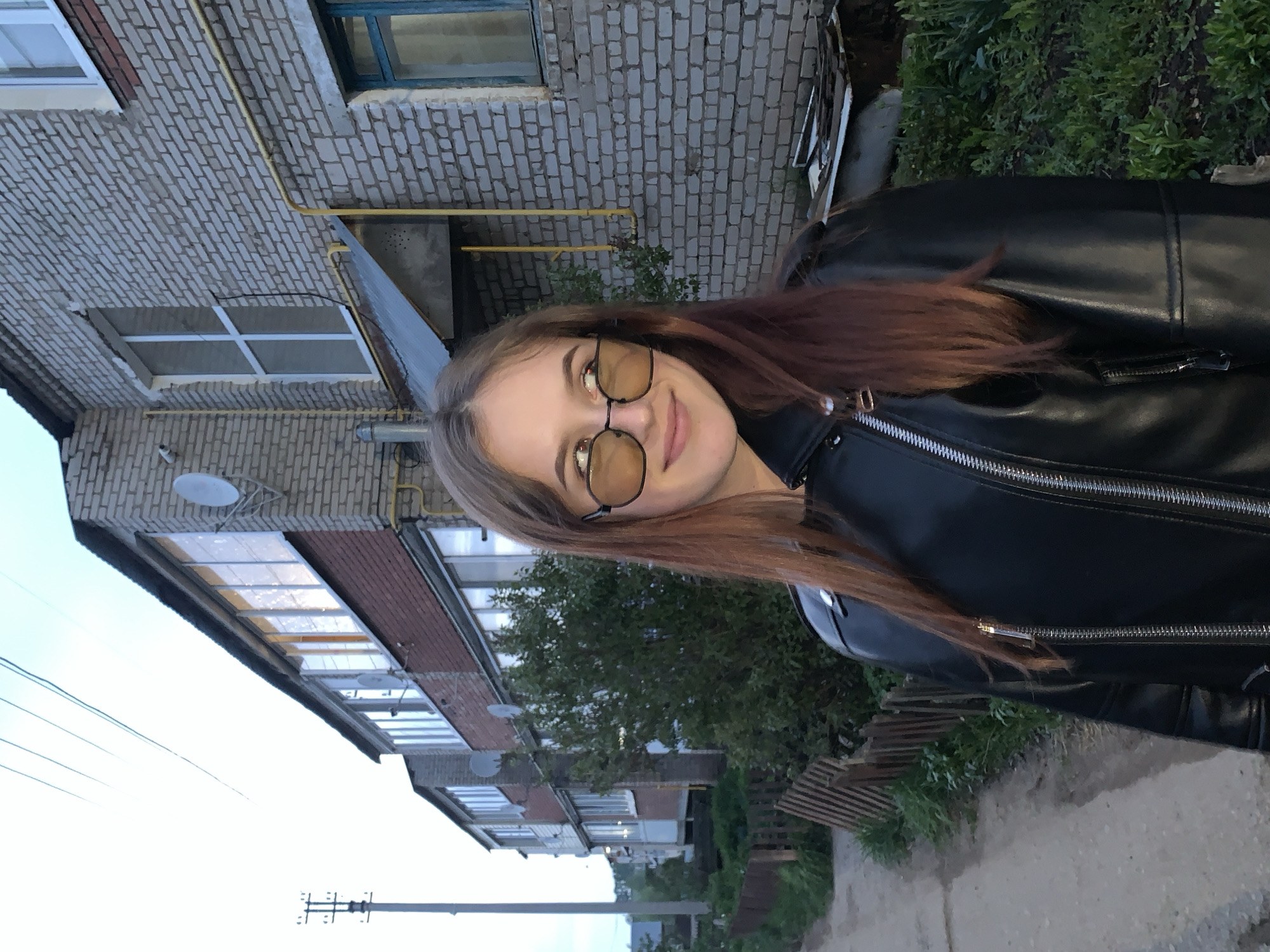 A young woman with light brown hair and glasses stands in front of a multi-story brick building, wearing a black leather jacket. The location appears to be in an urban area, possibly the city of Фоки, Russia, given the architectural style and surrounding environment. The background includes greenery and a street. The image is taken at an angle, with the building appearing to lean.