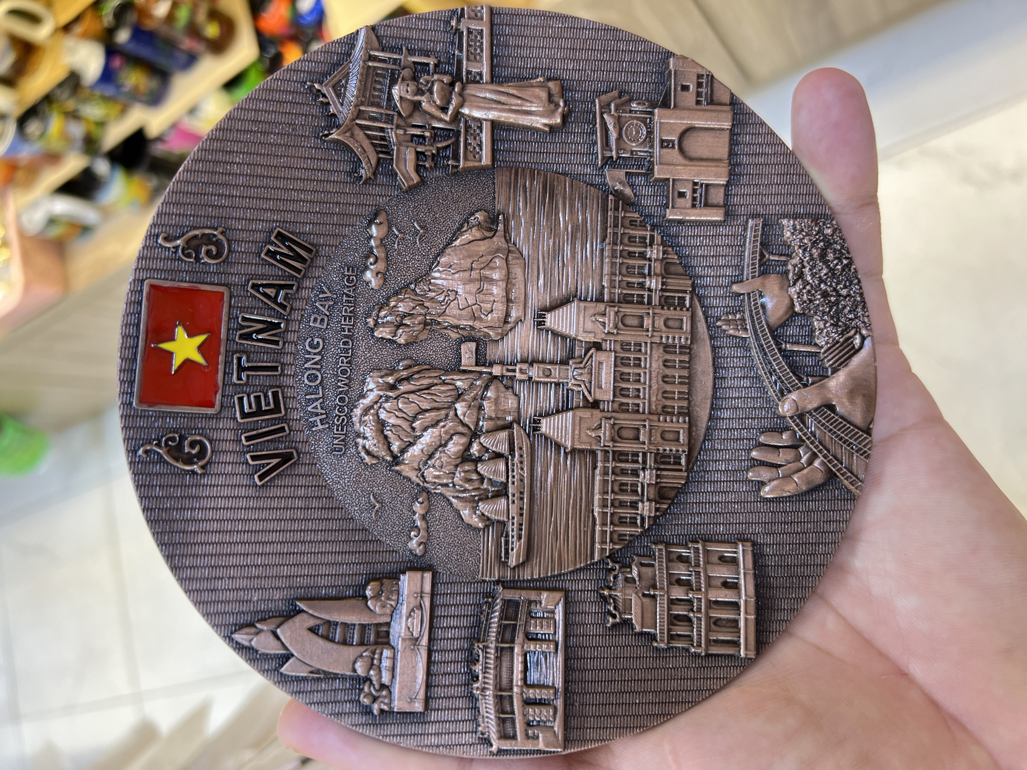 A circular, bronze-colored souvenir plate with intricate embossed details is being held by a person's hand. The plate features a prominent depiction of Vietnam's flag with a yellow star on a red background at the top, followed by the word "VIETNAM" in large letters. Below this, "HALONG BAY UNESCO WORLD HERITAGE" is inscribed. The central imagery includes stylized mountains, a boat, and various architectural structures. On the left side, there is an image of a unique tower, followed by a building resembling a temple or pagoda, and further down, another multi-story building. On the right, a figure of a woman in traditional Vietnamese attire stands beside a structure. At the bottom, a hand is shown holding a bridge-like structure, with other buildings and elements completing the scene. The background of the plate has a textured, ribbed pattern. The person's hand appears to be holding the plate from the bottom and left side, with skin texture visible. The environment in the background is out of focus, showing shelves with various items that appear to be souvenirs or merchandise in a retail setting. There is no identifiable element in the image that specifically indicates the location within the city of Nha Trang, Vietnam.