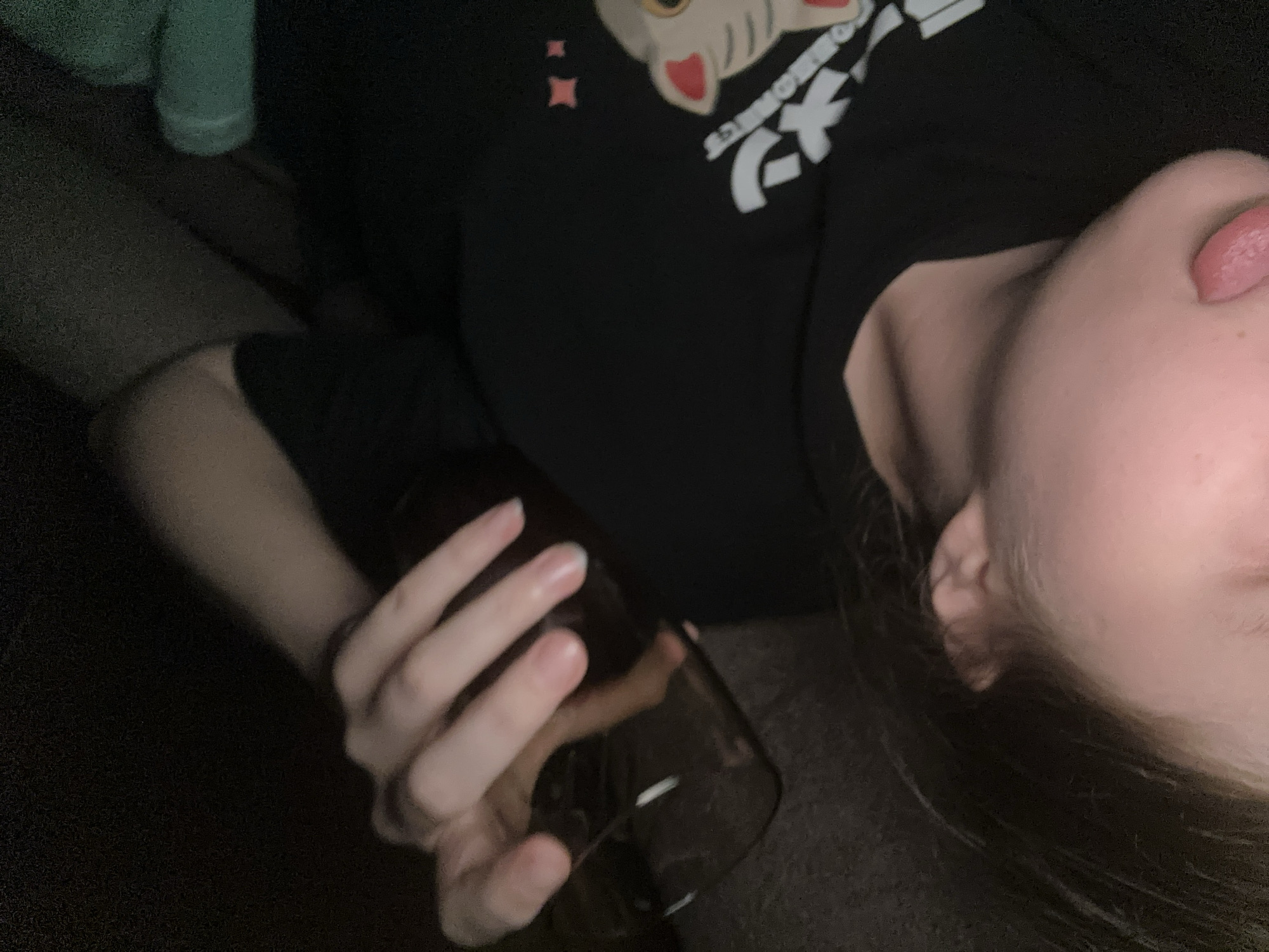 A person lies on their left side holding a glass bottle with their left hand. The person has fair skin, long brown hair, and is wearing a black t-shirt with a cat cartoon print and Japanese characters. The person's tongue is protruding from their mouth. The background is dark and indistinct, but appears to show a couch and an object wrapped in a green material. There are no clear indications of this being in Moscow, Russia.