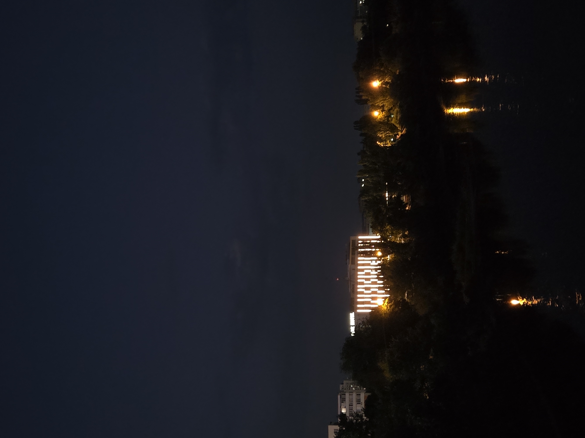 The image depicts a nighttime scene in Kitskany, Moldova, featuring a building illuminated by vertical stripes of light, likely part of its architectural design or a decorative lighting scheme. The building appears to be a hotel, as suggested by visible lettering that reads "PARK HOTEL". The structure is surrounded by dense foliage, forming a dark, silhouetted landscape. A body of dark water, possibly a river, occupies the lower portion of the frame, reflecting some of the artificial lights from the shore and the building. The sky is a deep, dark blue, suggesting late evening or night, with faint clouds visible. Several streetlights or outdoor lamps are also visible among the trees, casting pools of warm yellow light. The overall impression is a tranquil, nocturnal urban environment.