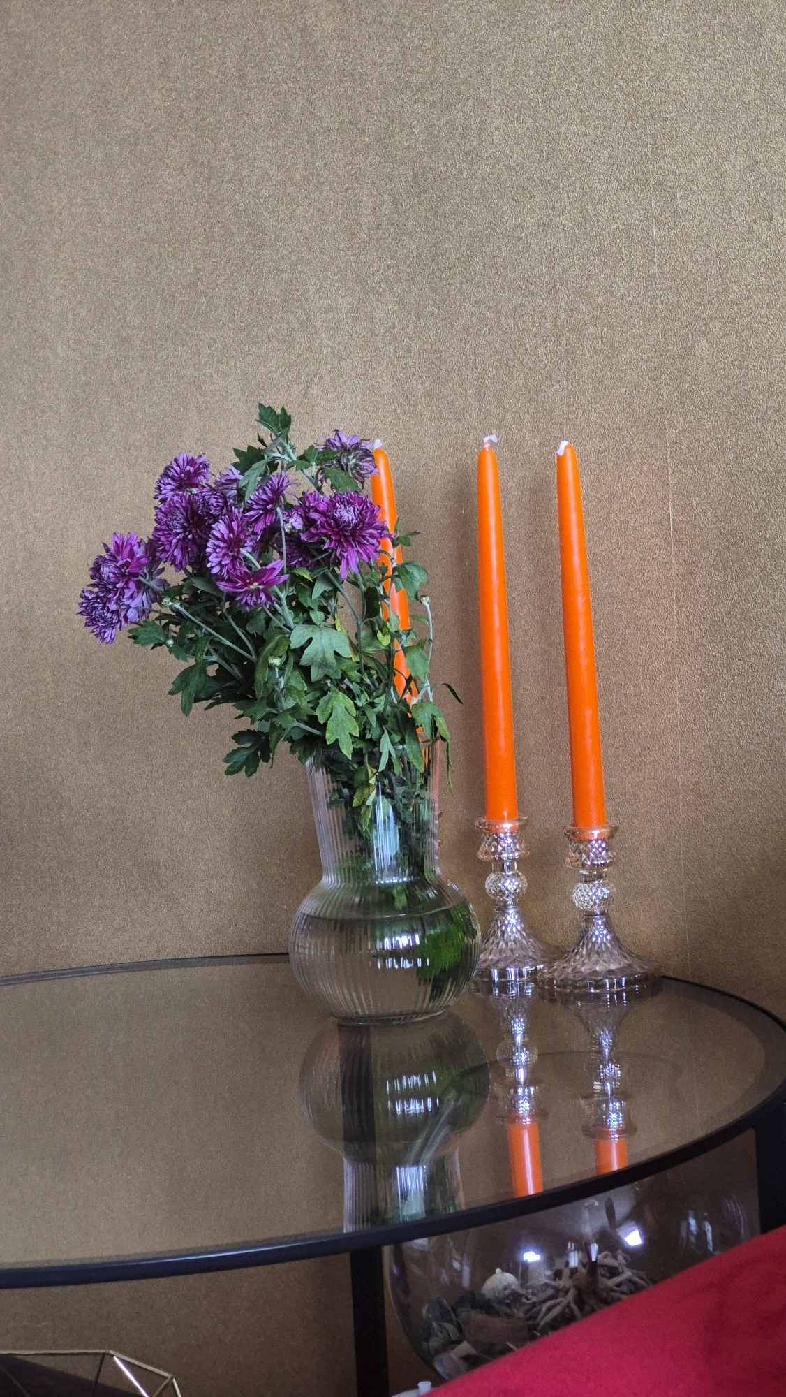 An arrangement of purple chrysanthemums in a clear glass vase sits on a reflective surface next to two orange tapered candles in ornate glass candleholders. The background is a textured, light brown wall. The scene appears to be indoors, likely on a table. There are no people visible. The overall impression is one of still life. The provided coordinates place this scene in Krauja, Latvia.