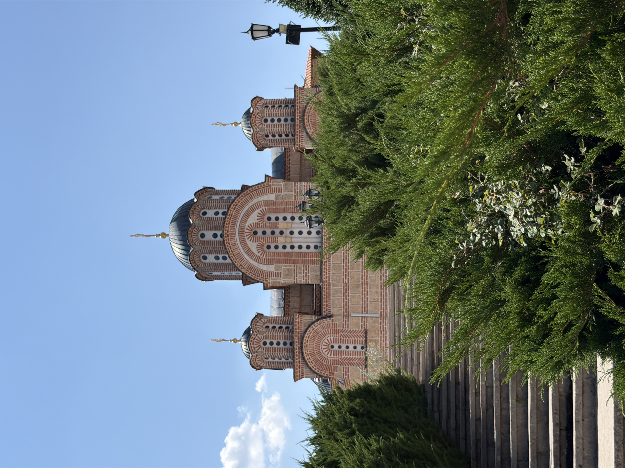 The image shows the exterior of the Hercegovačka Gračanica monastery, located on the hilltop overlooking the city of Trebinje, Bosnia and Herzegovina. The monastery features several domes with golden crosses, constructed with reddish bricks and stone. In the foreground, lush green bushes partially obscure a wide, stone staircase leading up to the monastery. A lamppost stands to the right of the monastery. The sky is a clear, bright blue with a few scattered clouds. There are no people visible in the image.